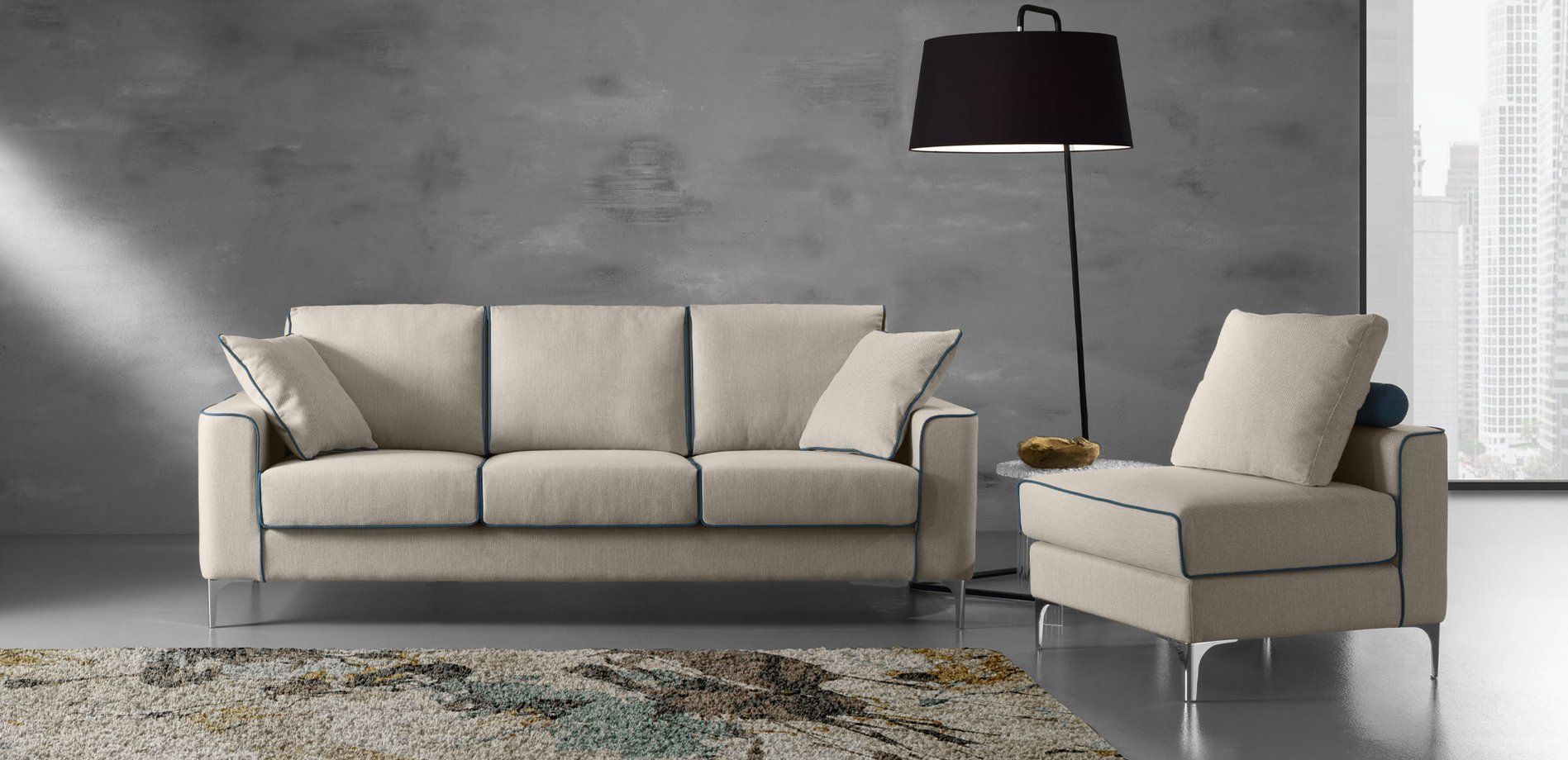 Divano e poltrona beige con finiture nere, lampada da terra e tappeto astratto contro una parete grigia.