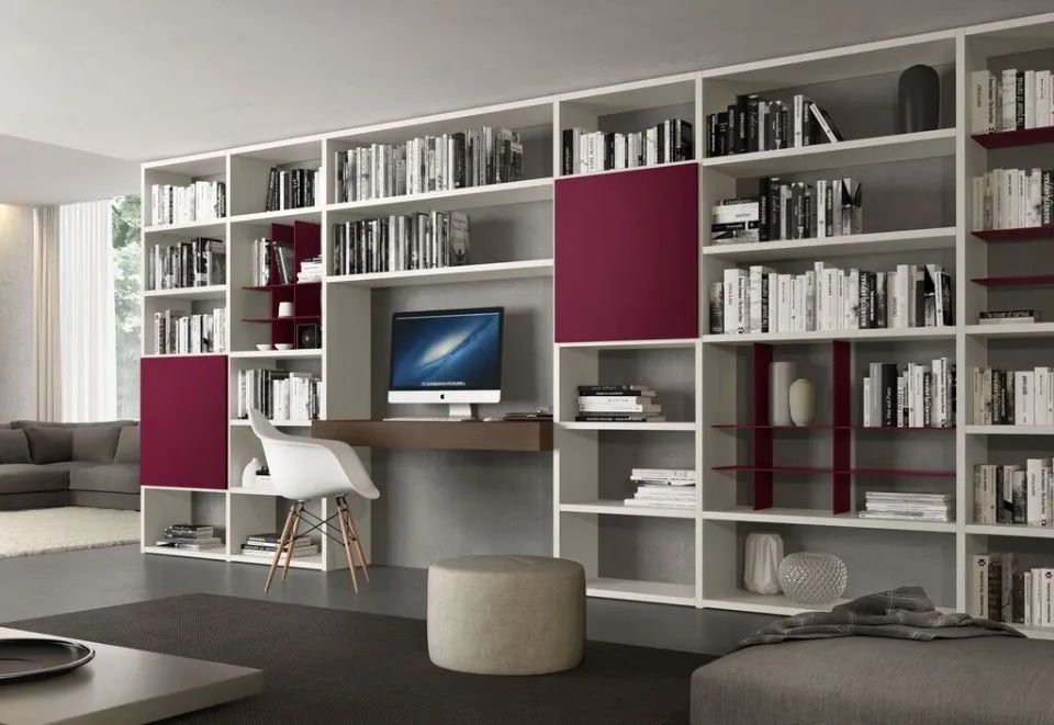 Ampia scaffalatura bianca con libri, scrivania, computer e sedia bianca. Sono presenti anche due pannelli rosso scuro.
