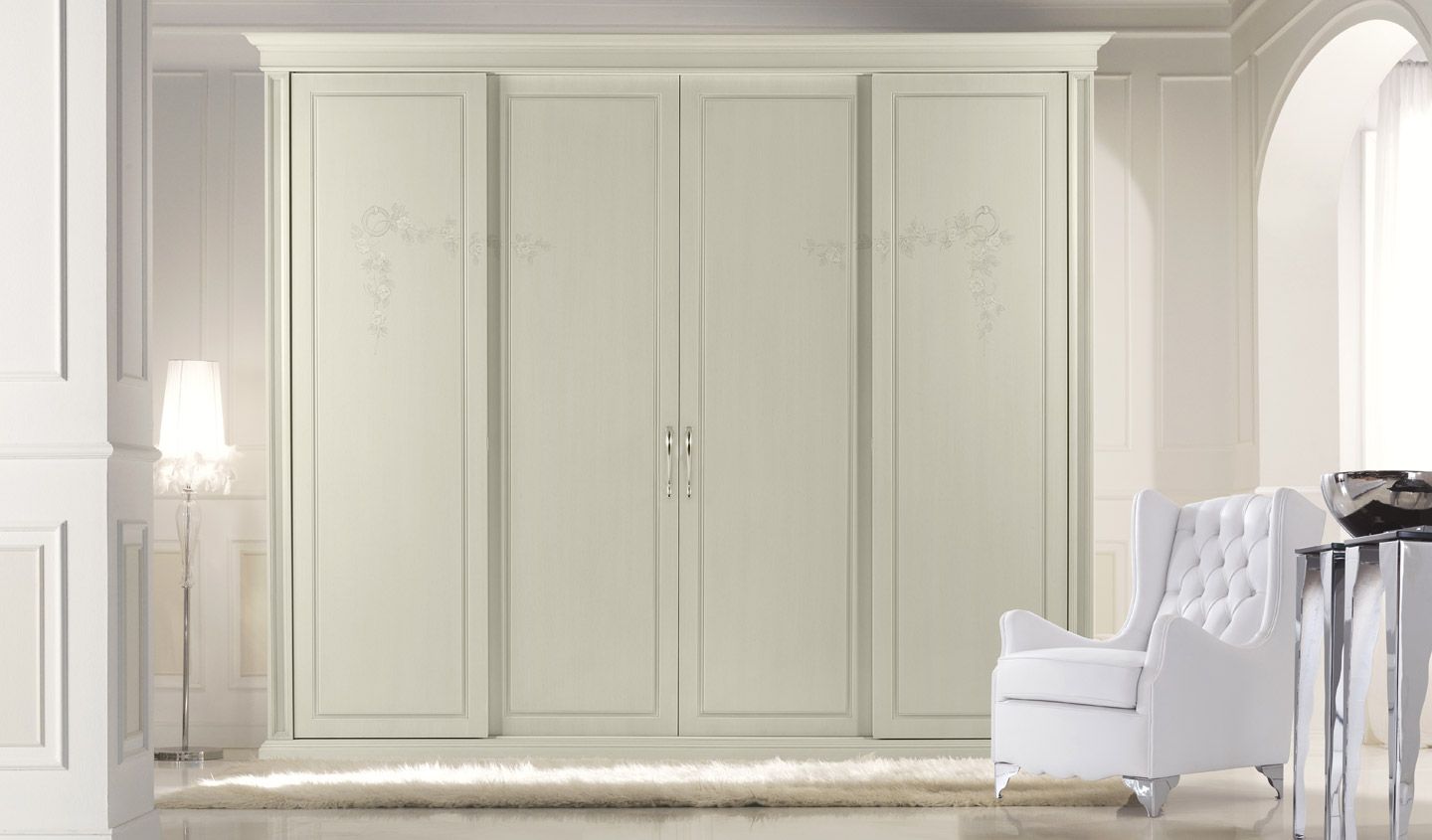Armadio color crema a quattro ante in una stanza bianca. Nelle vicinanze si trovano una poltrona bianca imbottita e un tavolo.