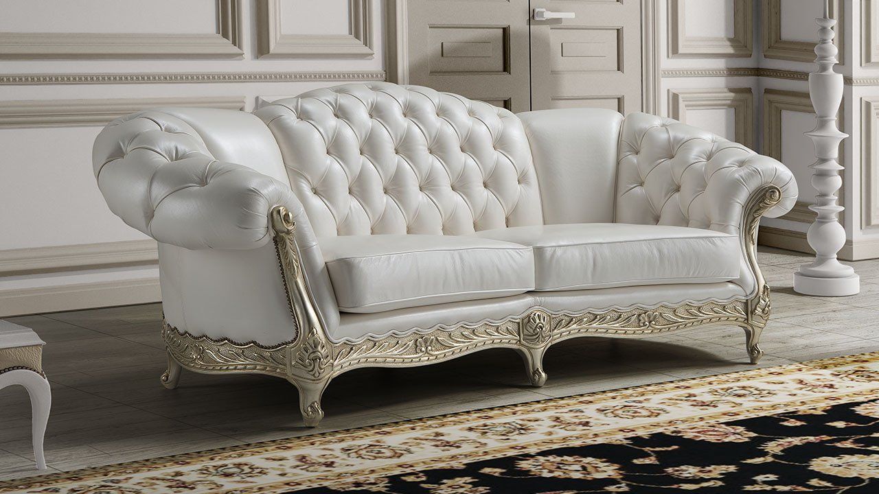 Divano bianco imbottito con rifiniture dorate elaborate, in una stanza con un tappeto a fantasia e pareti dai colori chiari.