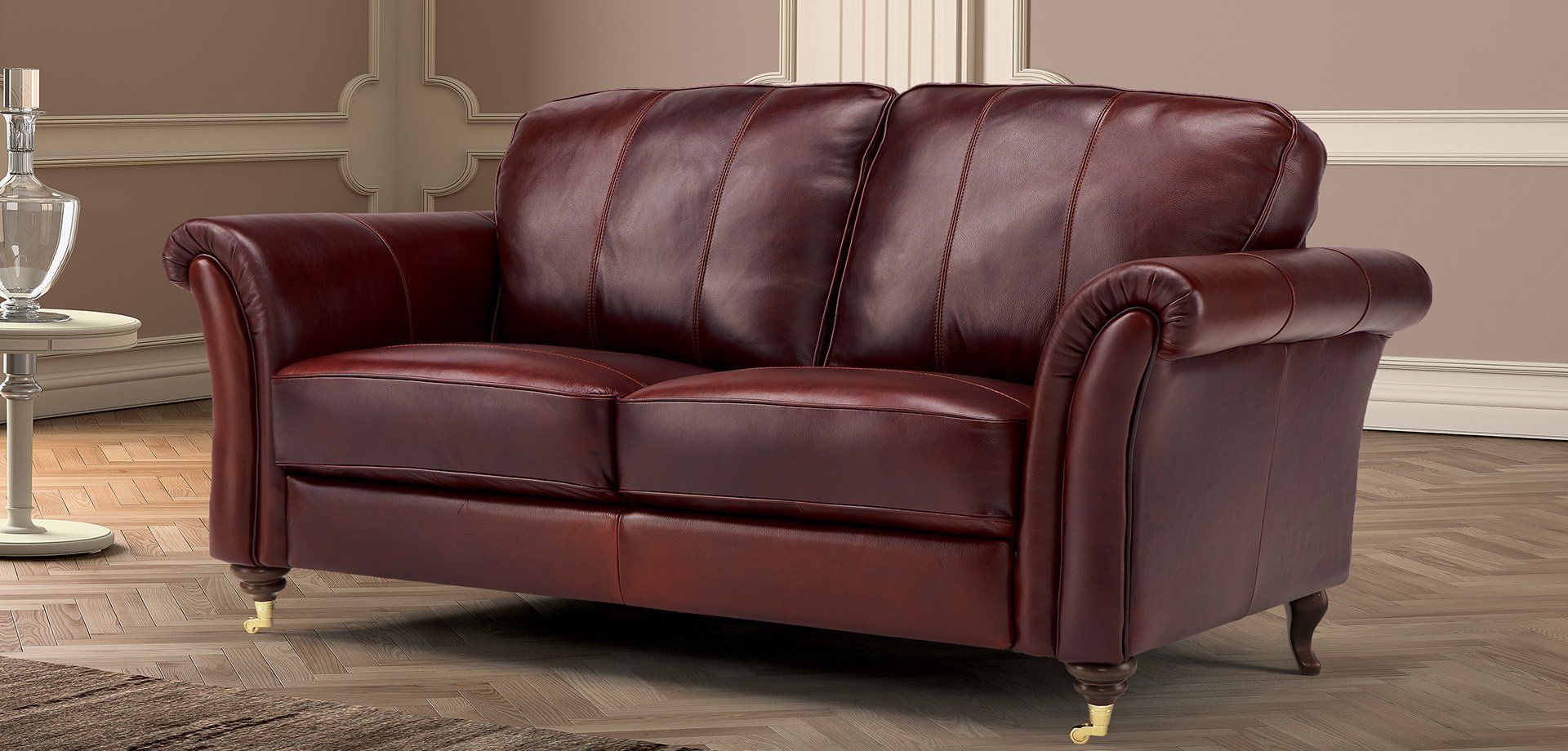 Divano in pelle bordeaux con braccioli arrotolati, borchie e piedini in ottone in una stanza con pareti chiare.