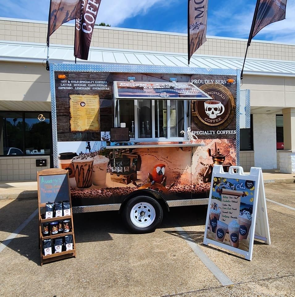 A branded mobile coffee trailer parked in a lot, with a menu board, coffee bags on display, and a sign for iced drinks.