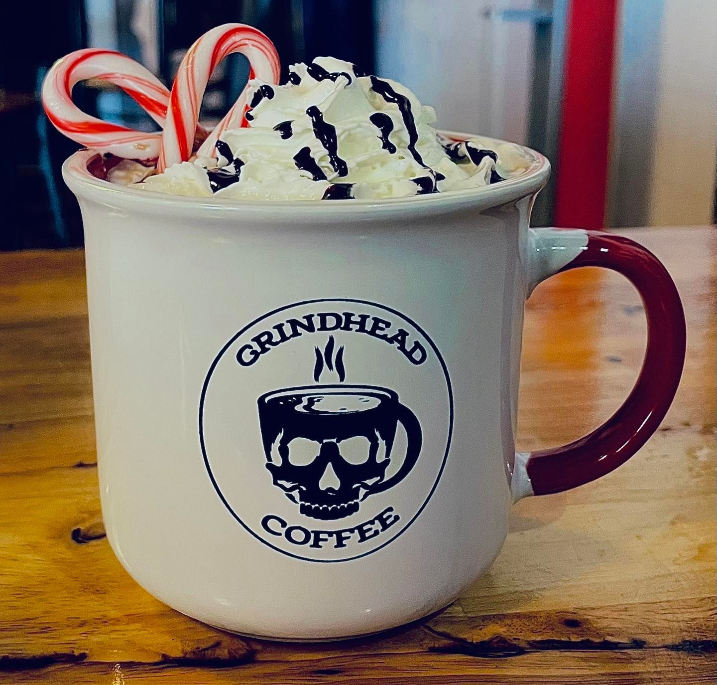 A Grindhead Coffee mug filled with hot cocoa, topped with whipped cream, chocolate drizzle, and a candy cane.