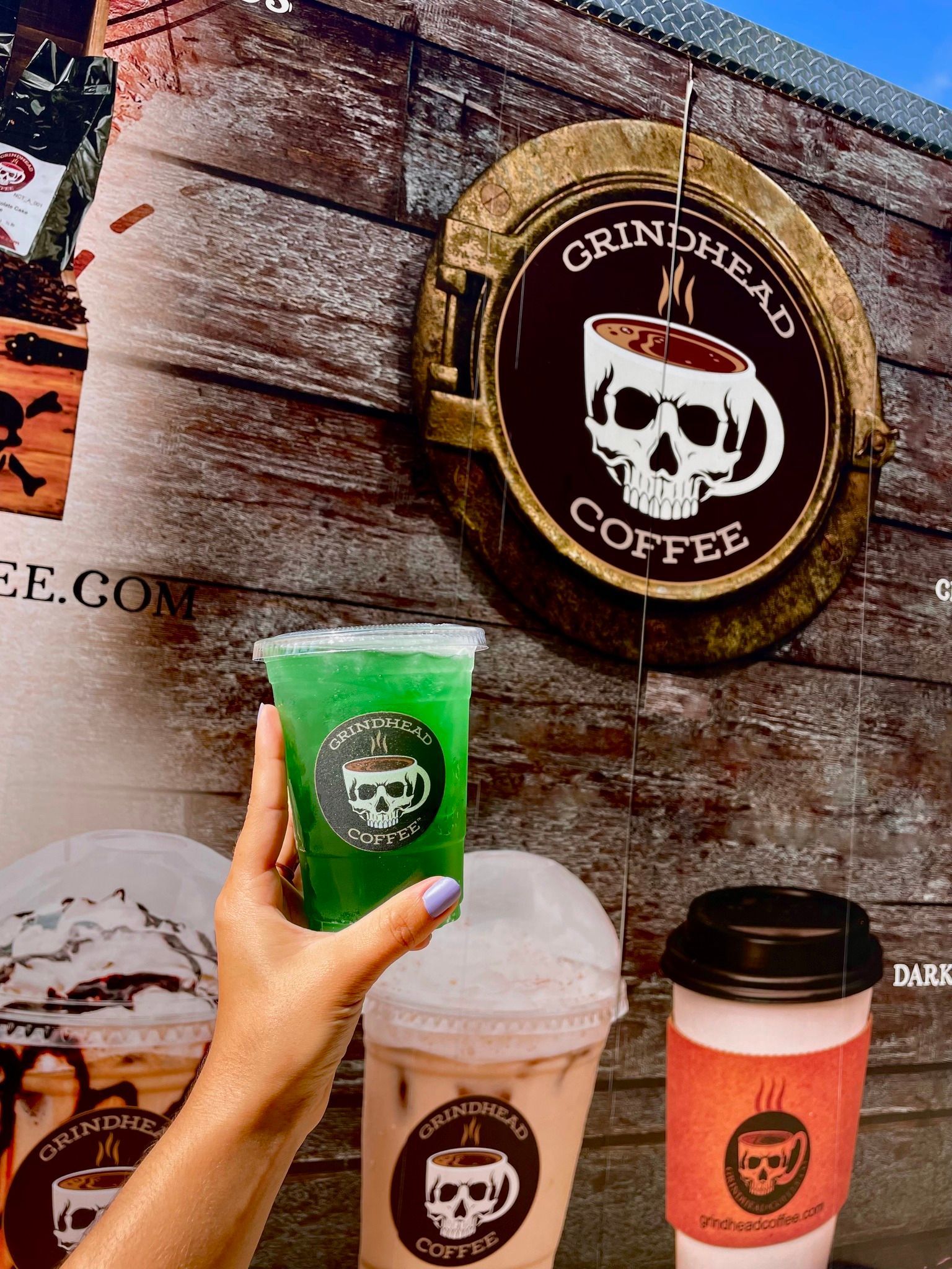 A hand holds a bright green drink in front of a Grindhead Coffee sign, with two other beverages on a surface below.
