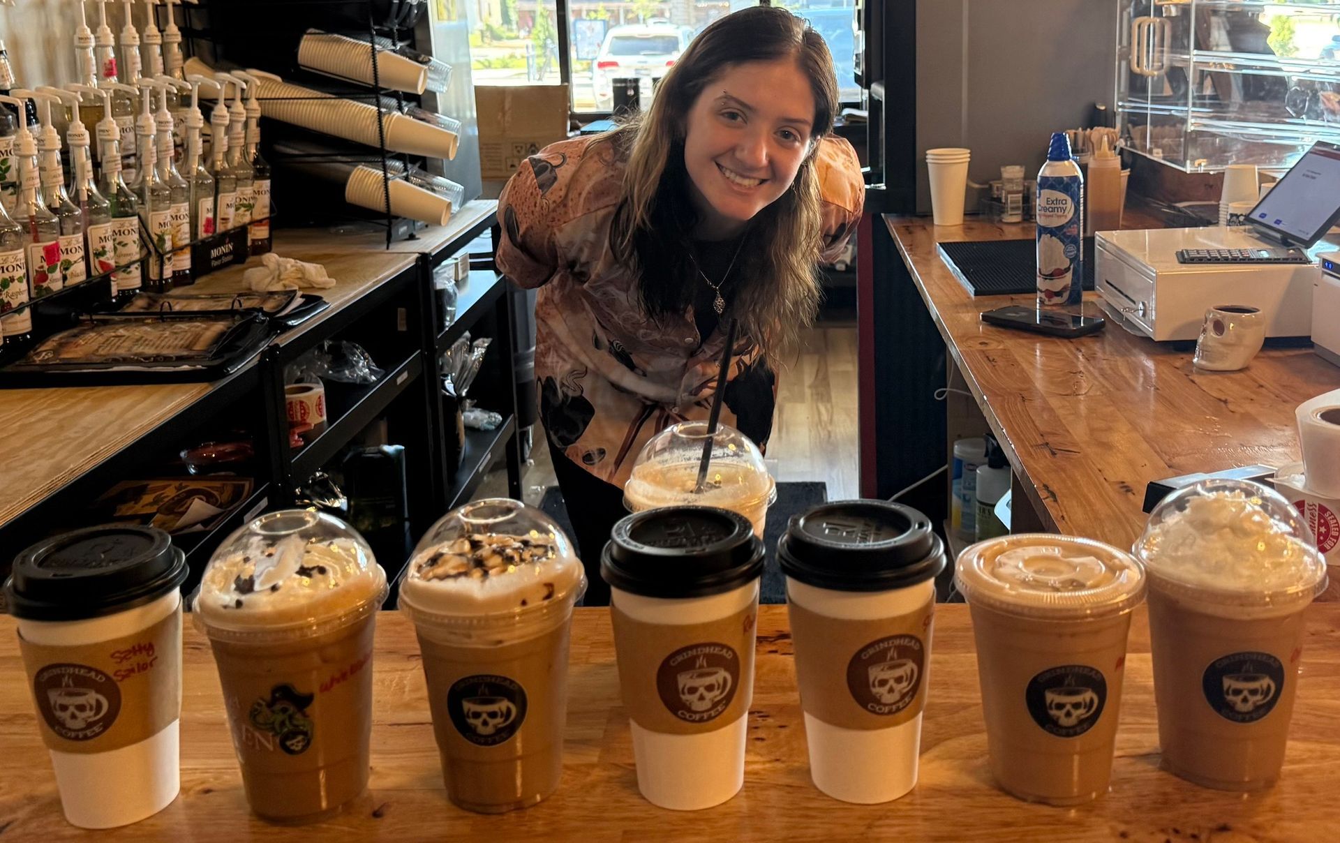 A smiling staff member stands behind a wooden counter lined with seven varied coffee drinks in a cafe.