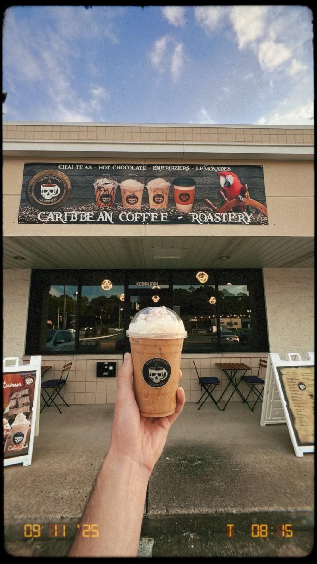A person holds a whipped cream-topped iced coffee in front of a Caribbean Coffee Roastery shop storefront.