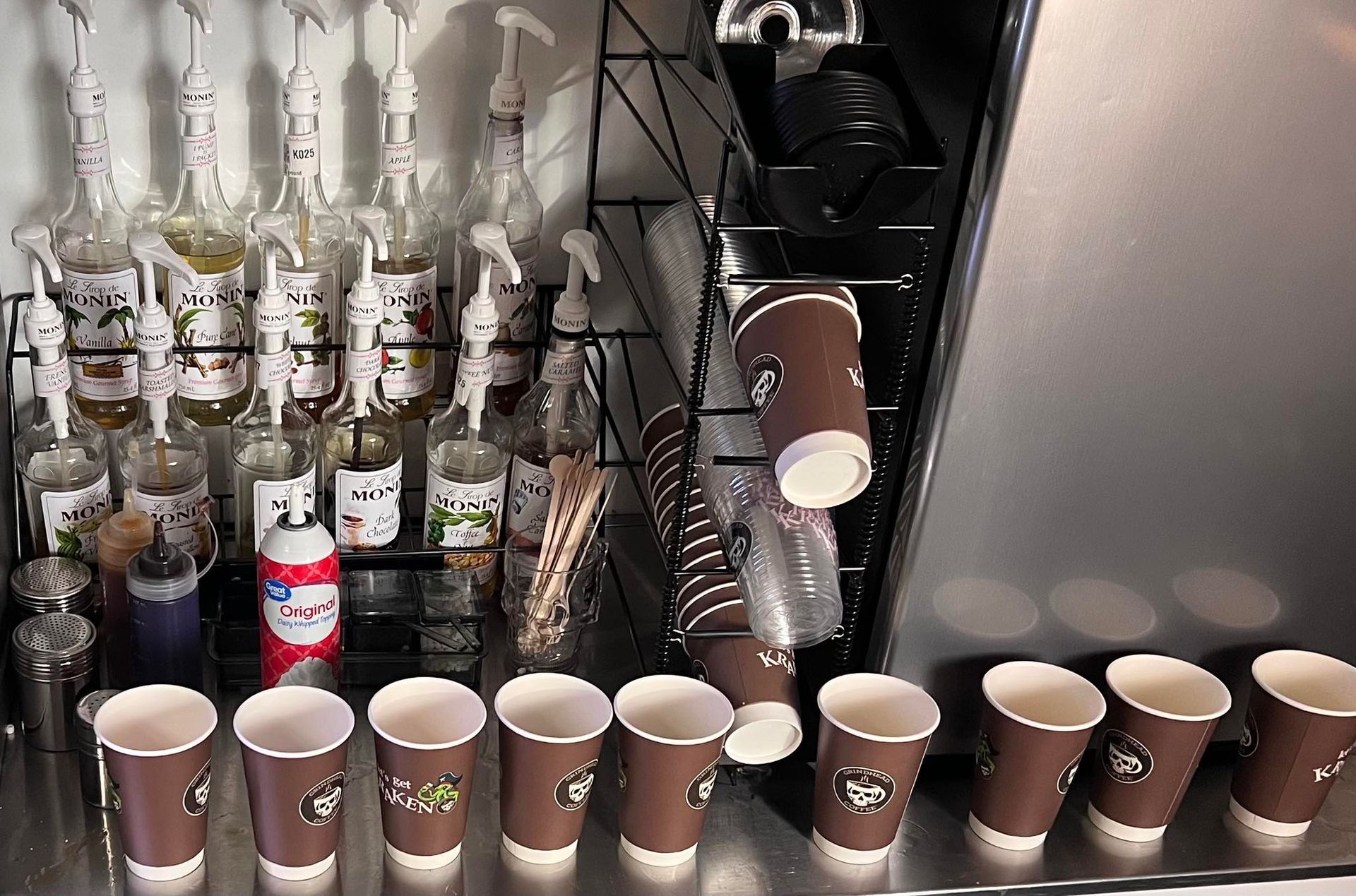 A coffee station featuring rows of brown paper cups, syrup bottles on a rack, and a silver espresso machine.