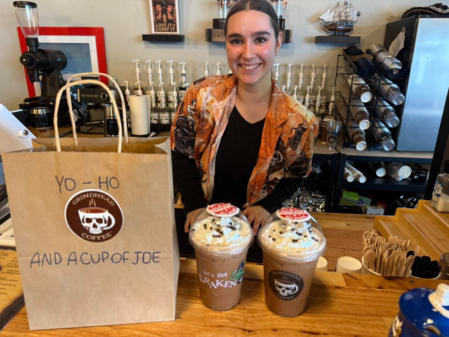 A smiling barista stands behind a counter with two iced coffee drinks and a paper bag labeled 