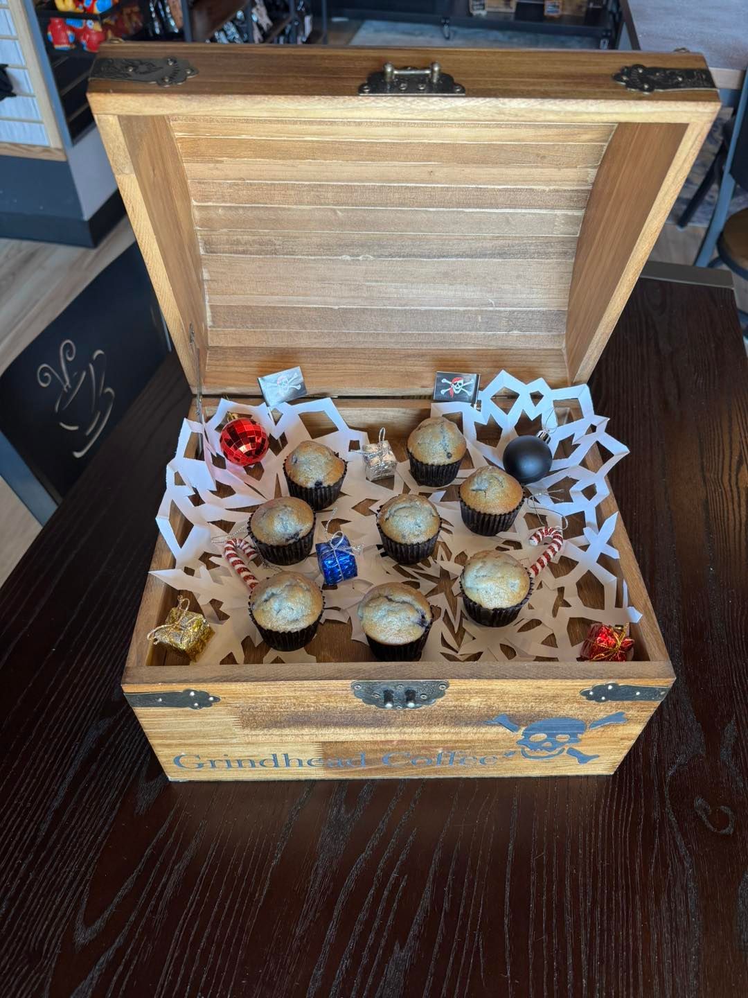 An open wooden chest contains mini muffins arranged on a white decorative paper doily with small festive ornaments.