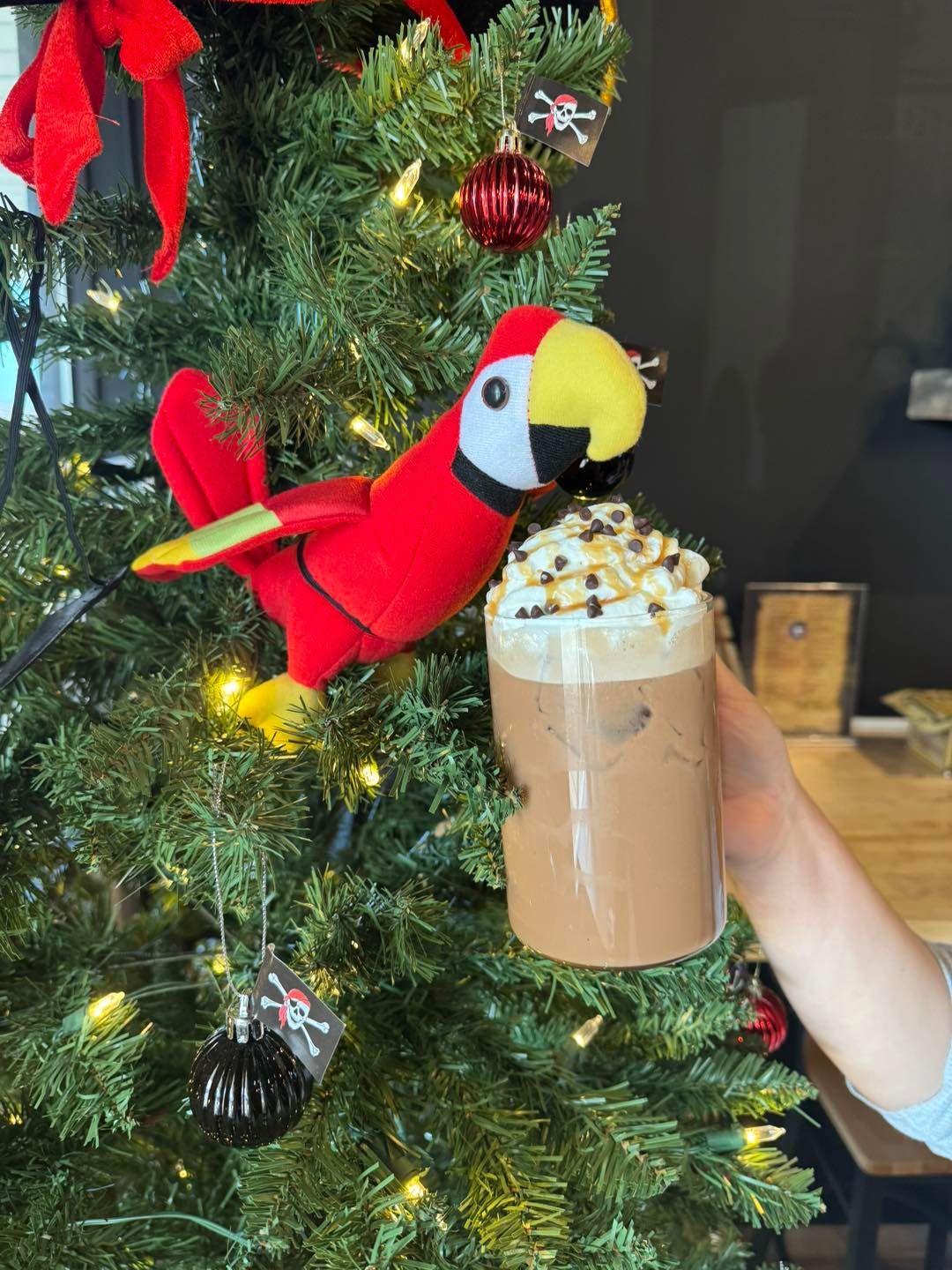 A hand holds a tall glass of chocolate coffee with whipped cream next to a parrot plush on a decorated Christmas tree.
