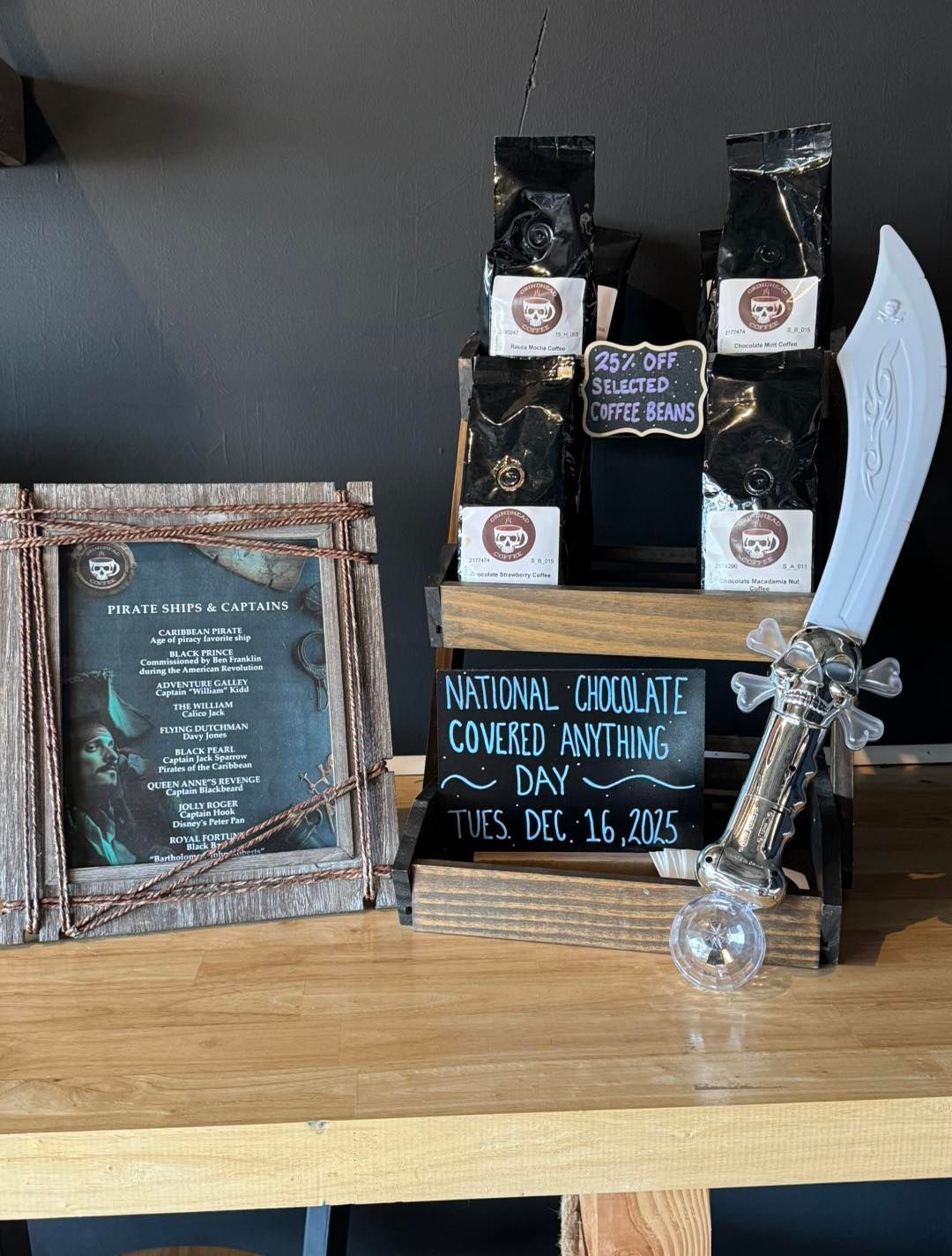 A wooden display on a table with bags of coffee beans, a decorative pirate sword, and signs for a special event.