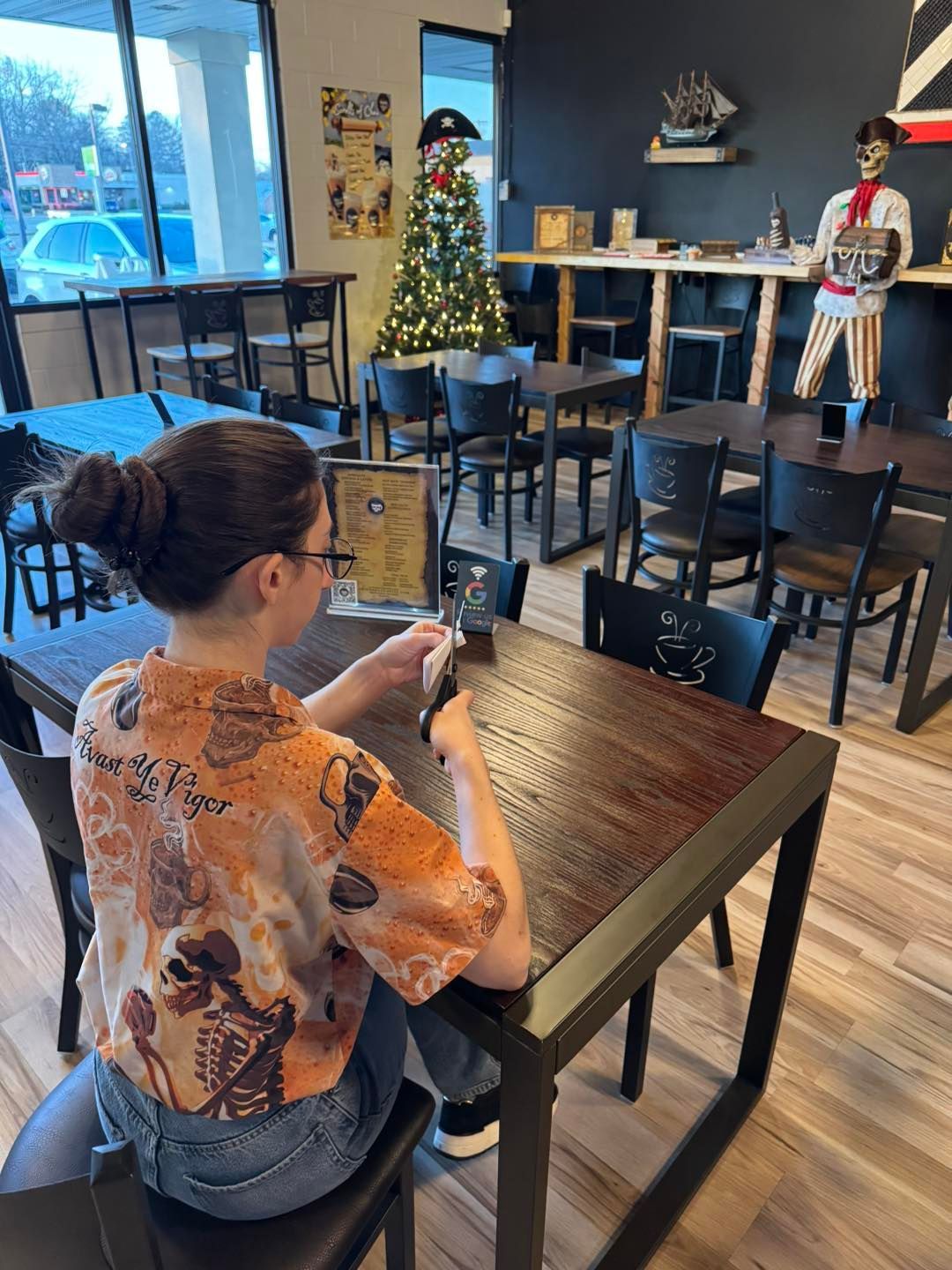A person in an orange patterned shirt sits at a dark wood table in a cafe decorated with a pirate theme and a tree.