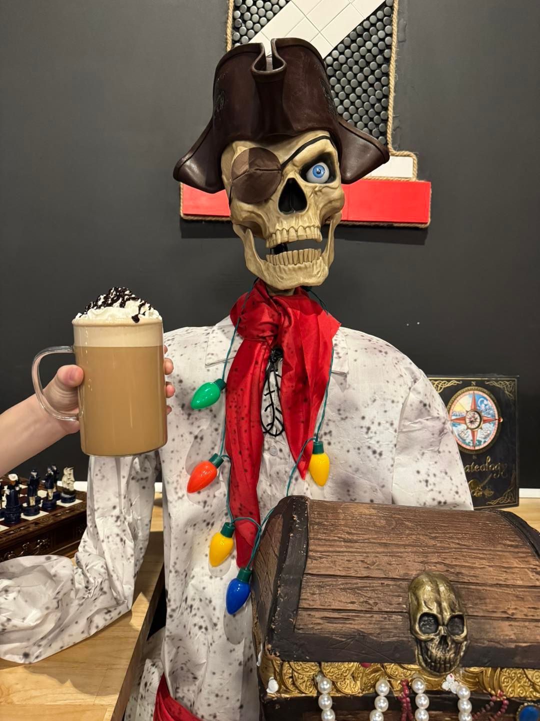 A pirate-themed skeleton in a tricorn hat and red scarf holds a latte, standing behind a chest with a skull emblem.