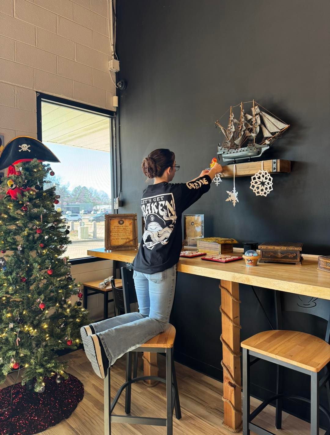A person kneels on a bar stool to reach a model ship on a wall shelf in a room decorated with a Christmas tree.