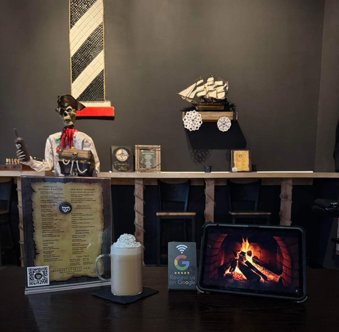 A pirate-themed coffee shop counter with a skeleton, a menu, a mug with whipped cream, and a tablet showing a fireplace.
