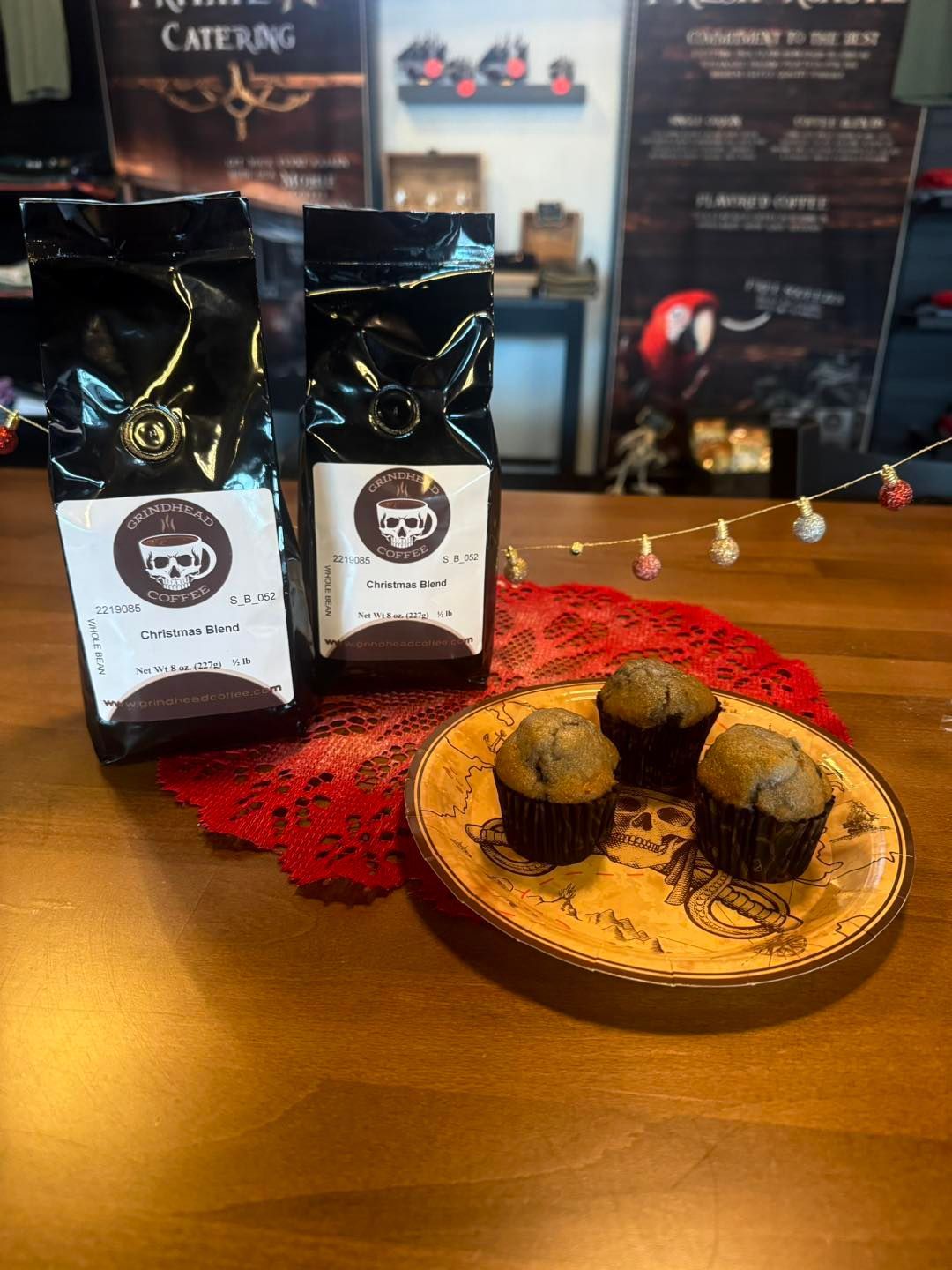 Two black coffee bags with skull logos sit behind a patterned plate holding three small chocolate muffins on a table.