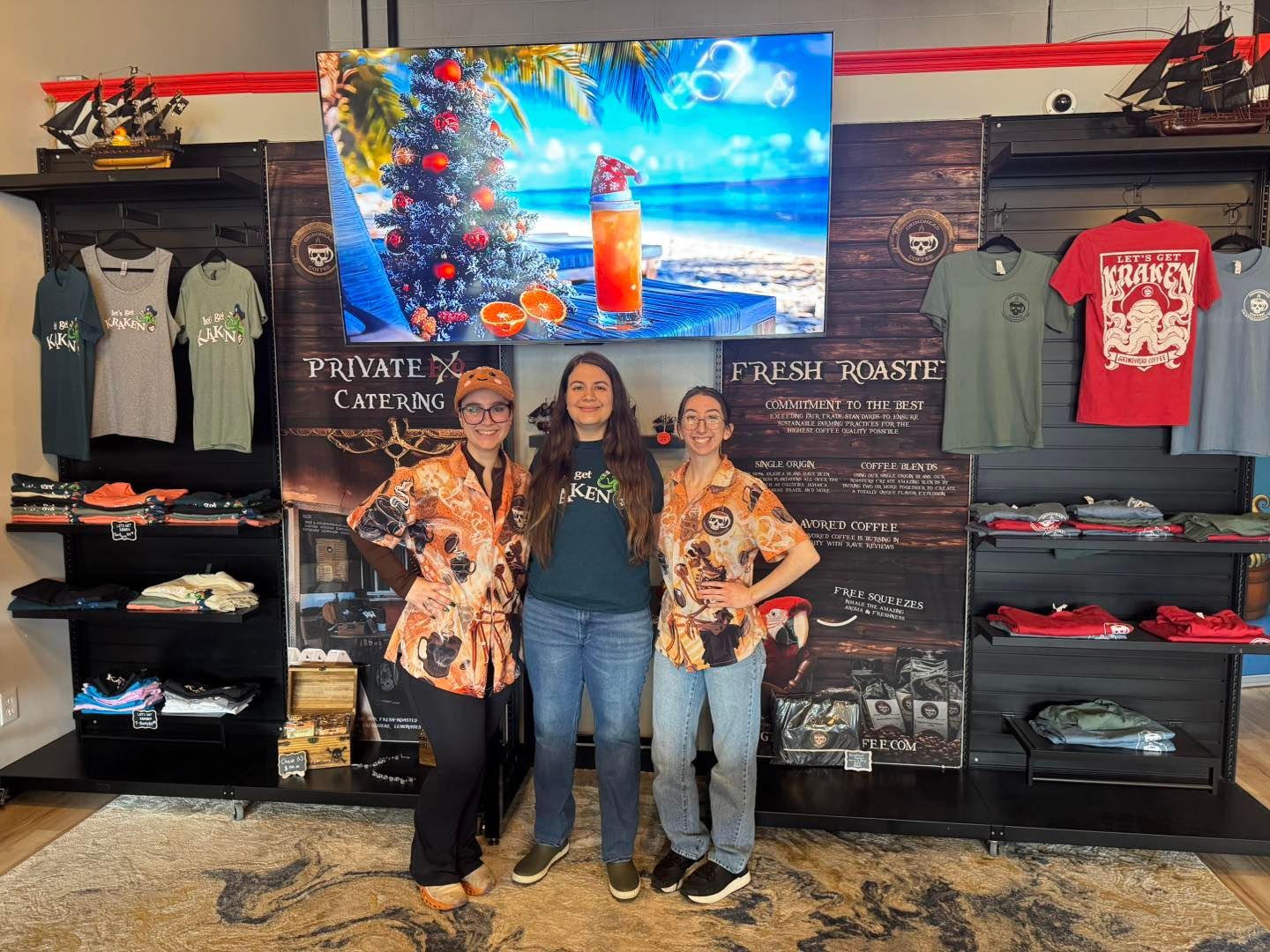 Three staff members stand in a retail store in front of clothing displays and a television screen showing a beach scene.