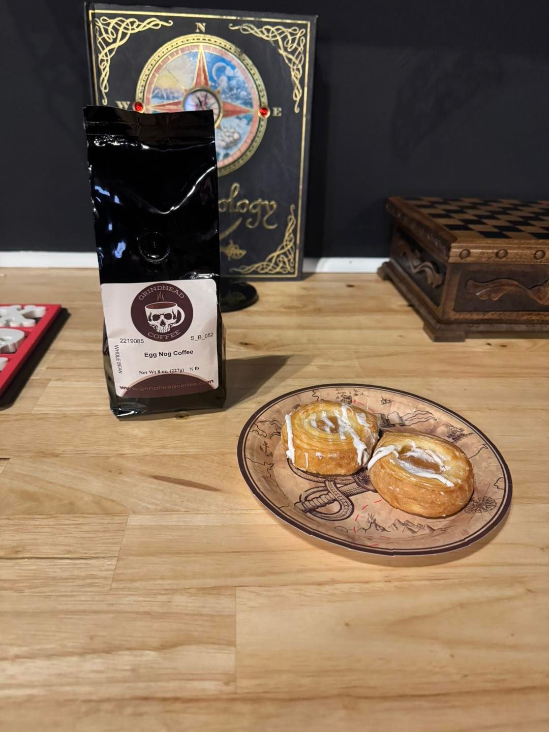 A bag of skull-logo coffee next to two small, glazed pastries on a patterned plate, set on a wooden table.