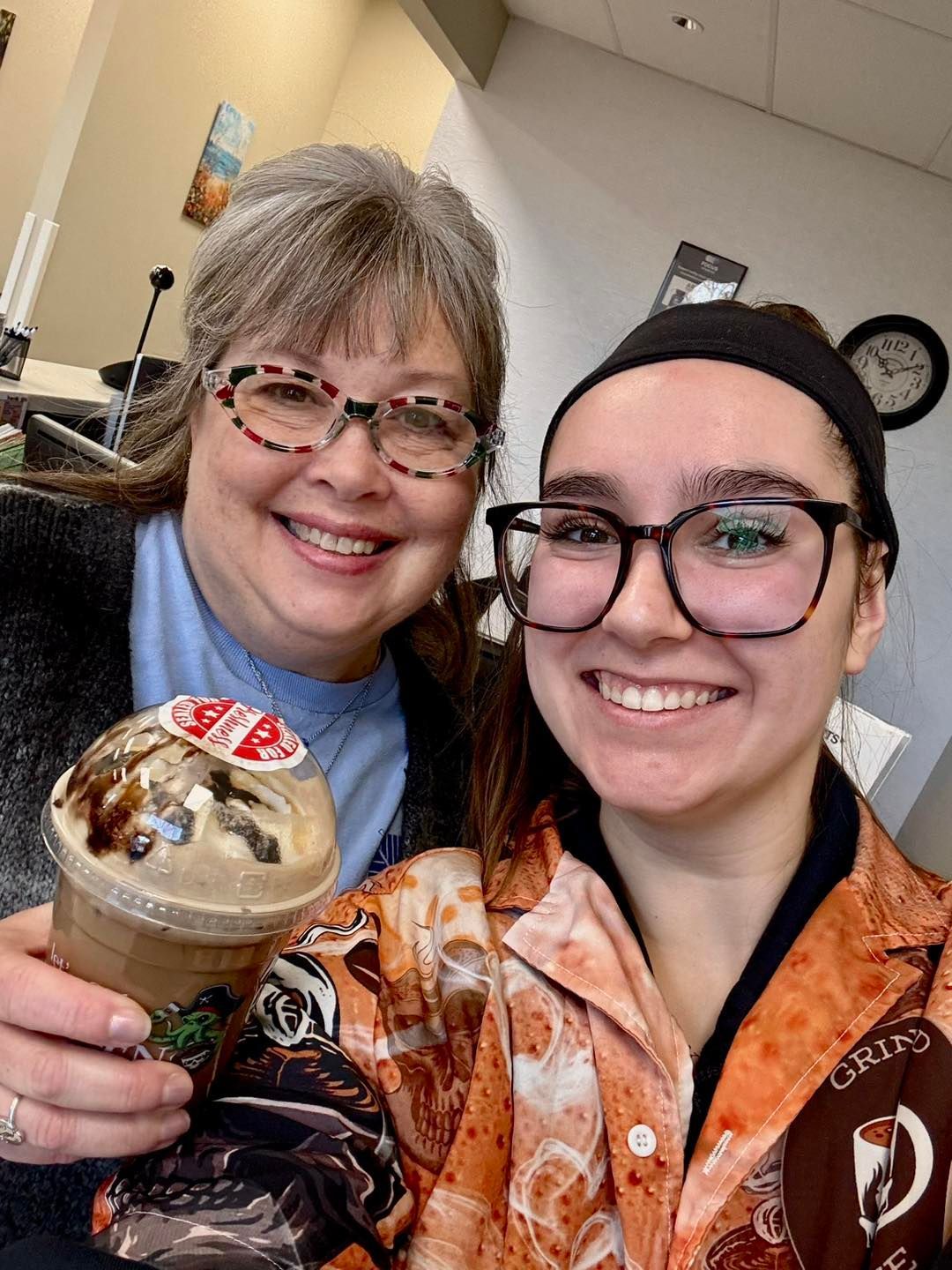 Two smiling people wearing glasses pose for a selfie, one holding a plastic cup of iced coffee in an indoor setting.