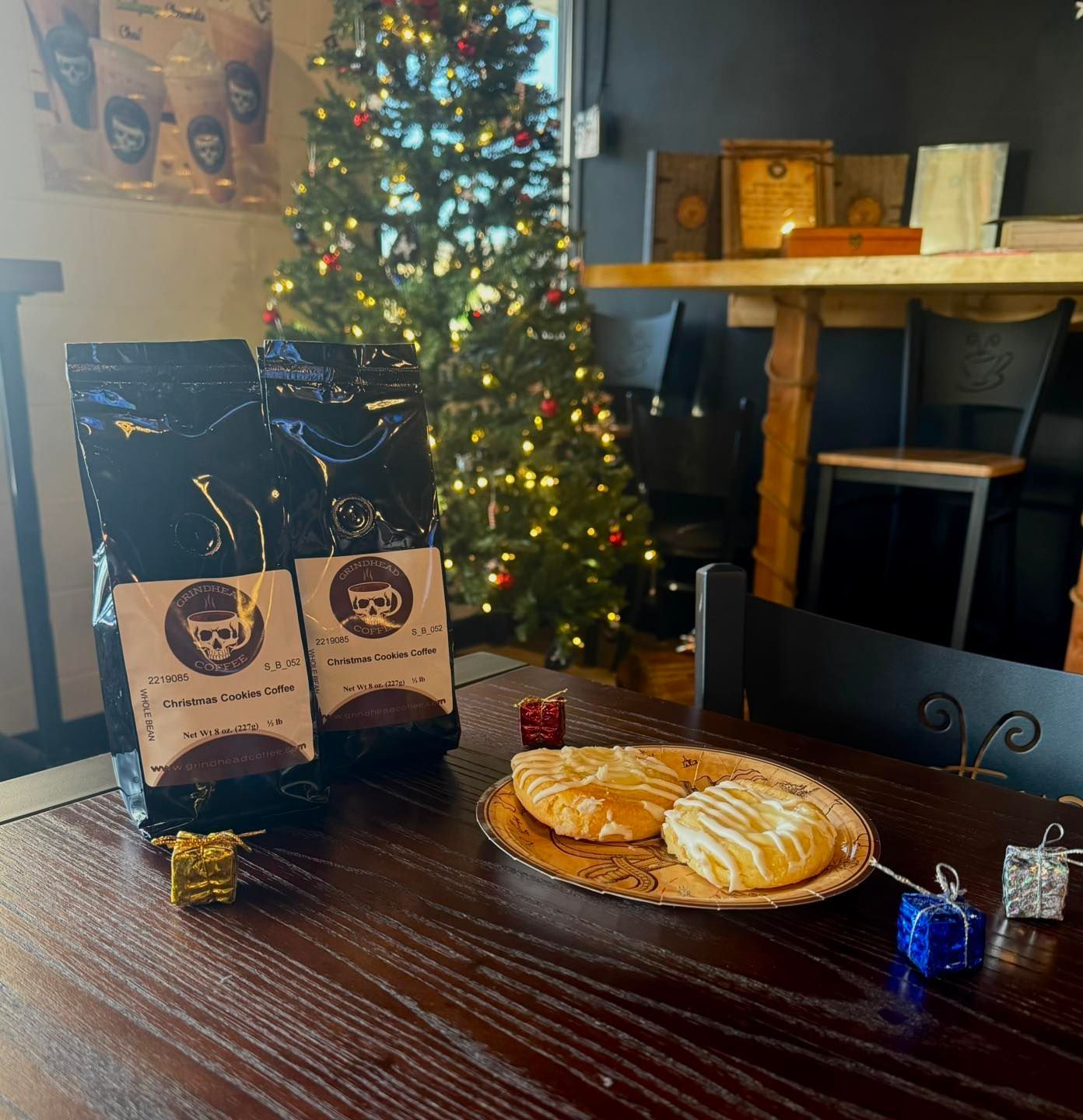 Two coffee bags and a plate with two pastries sit on a dark wooden table near a decorated Christmas tree.