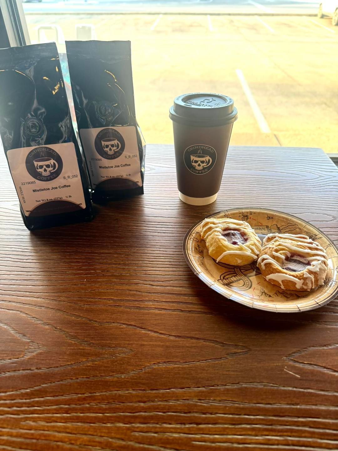 Two bags of coffee, a takeaway cup, and a plate with two small pastries sit on a wooden table.