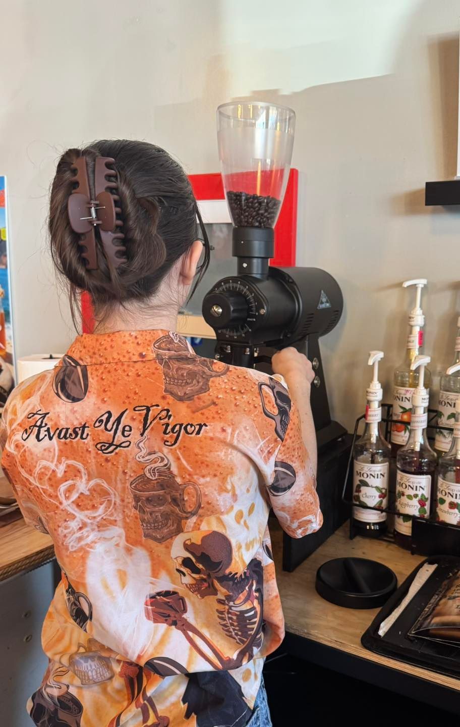 A barista wearing an orange patterned shirt uses an industrial coffee grinder in a cafe setting with syrup bottles nearby.