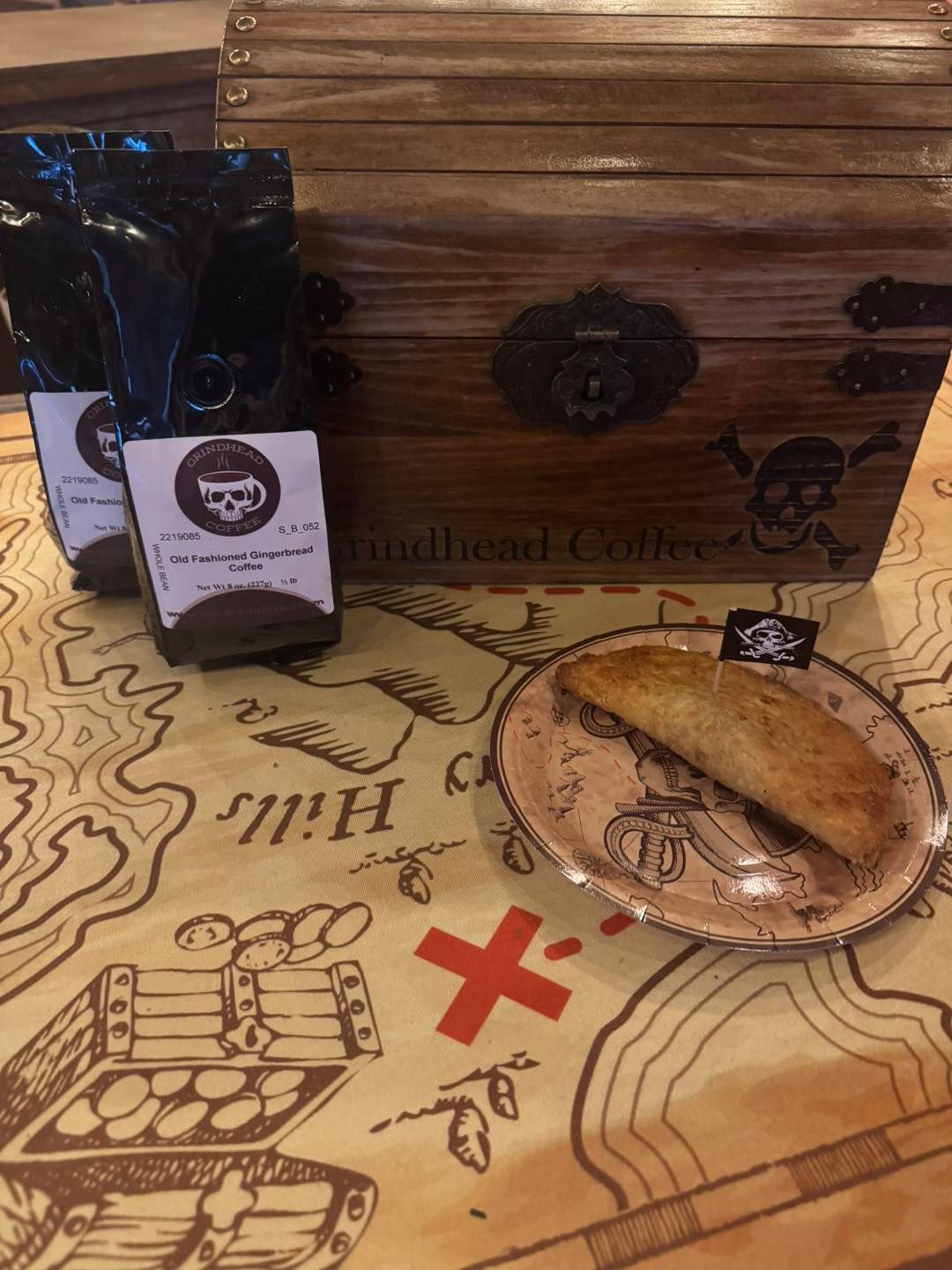 Two bags of Skullhead Coffee, a wooden chest, and a pastry on a plate placed on a table with a treasure map pattern.