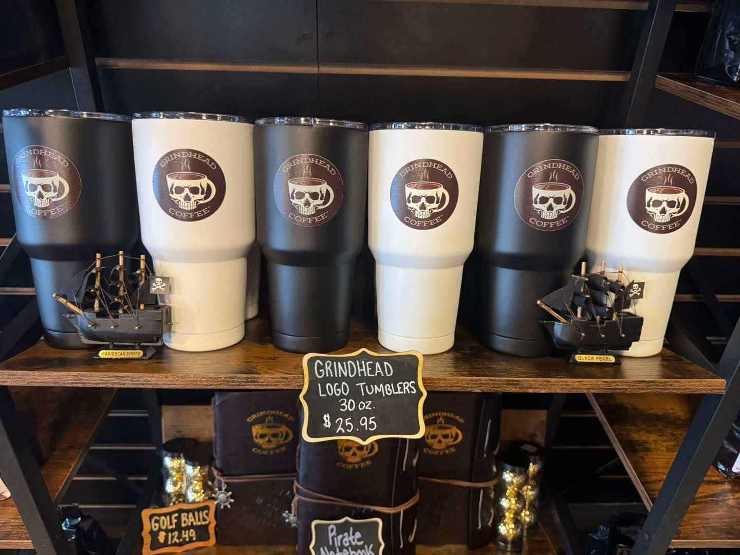 Black and white tumblers with skull logos on a shelf, with a small sign for Grindhead Logo Tumblers priced at $25.95.