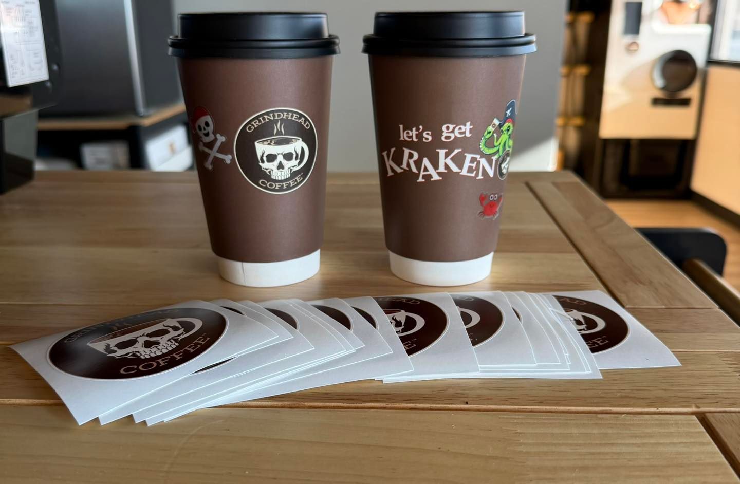 Two brown coffee cups with skull and Kraken logos sit on a wooden table, surrounded by several stickers.