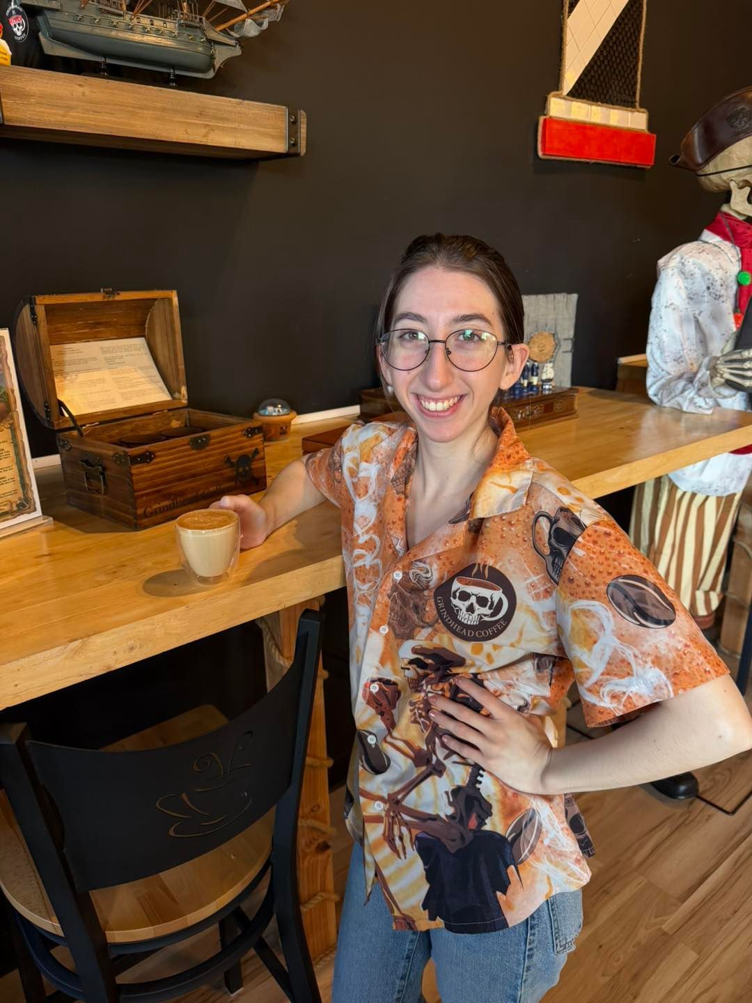 A smiling person wearing a patterned button-down shirt stands in a pirate-themed cafe, holding a coffee mug.