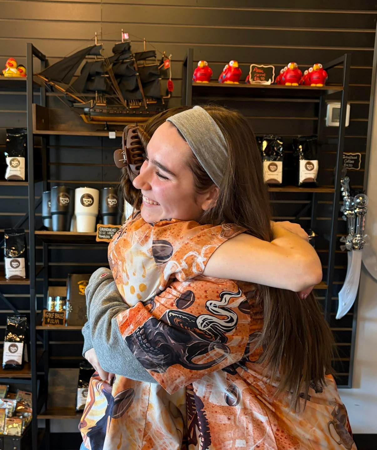 A person wearing a grey headband and a patterned shirt smiles while embracing another person inside a retail shop.