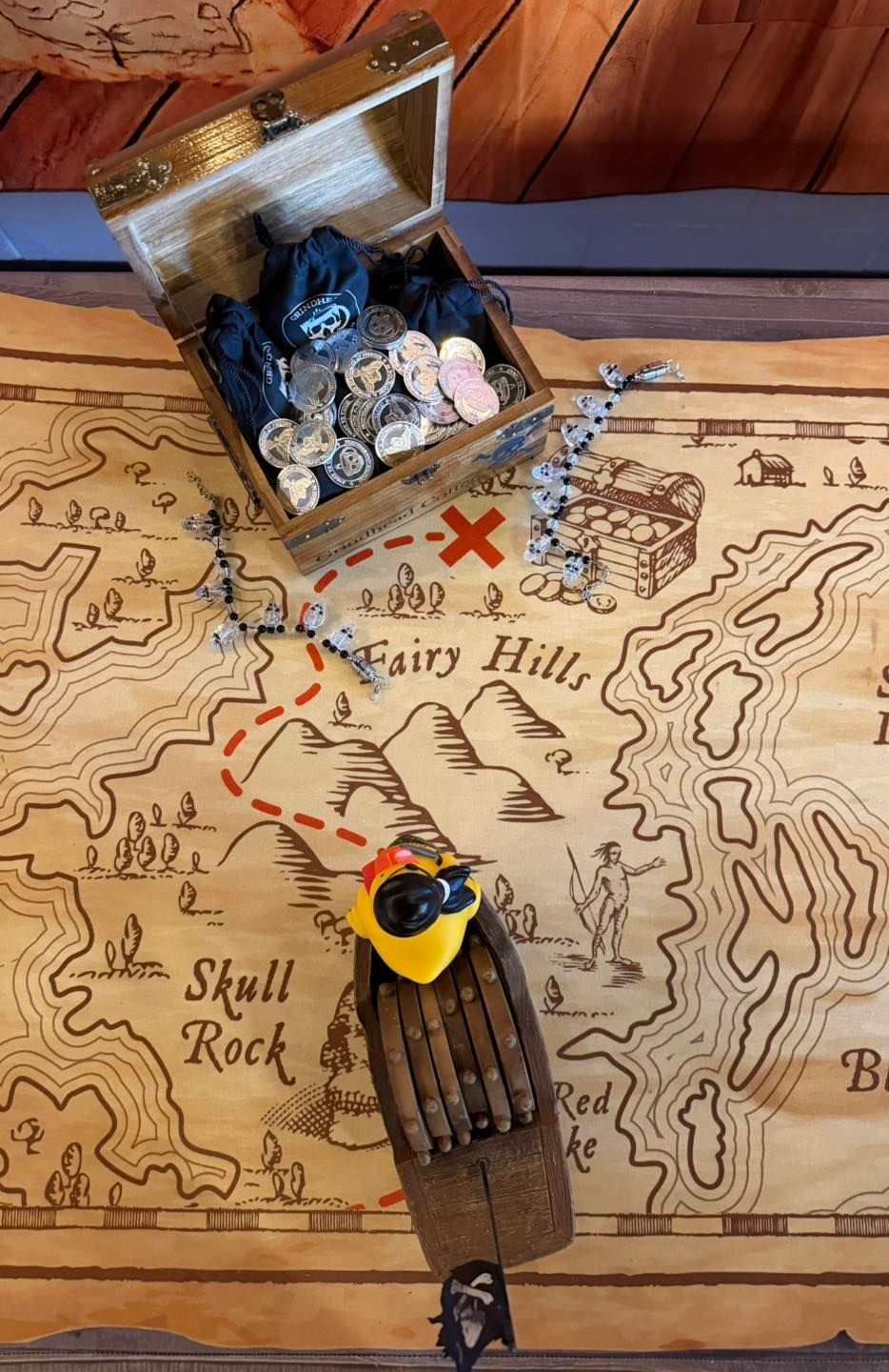 A treasure chest filled with coins sits on a treasure map marked with a red X, next to a small toy boat with a yellow bird.