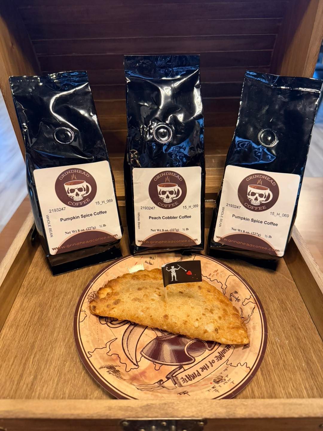 Three bags of Black Rifle Coffee Company coffee stand behind a fried hand pie on a decorative plate inside a wooden crate.