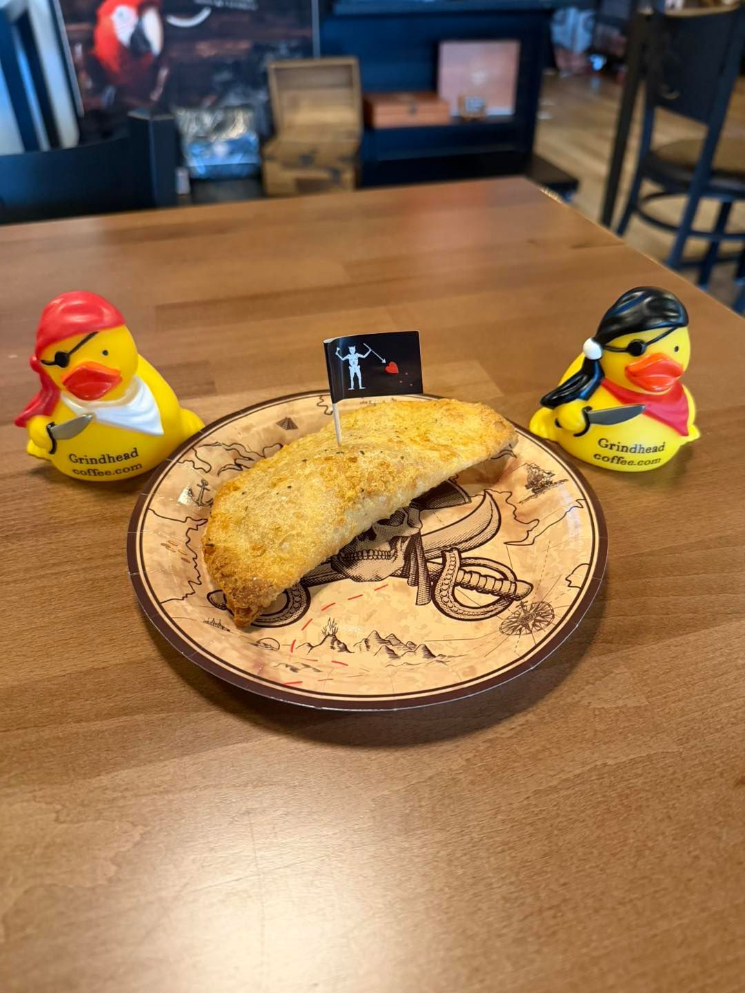 A breaded pastry with a pirate flag sits on a patterned plate between two pirate-themed rubber ducks on a wood table.