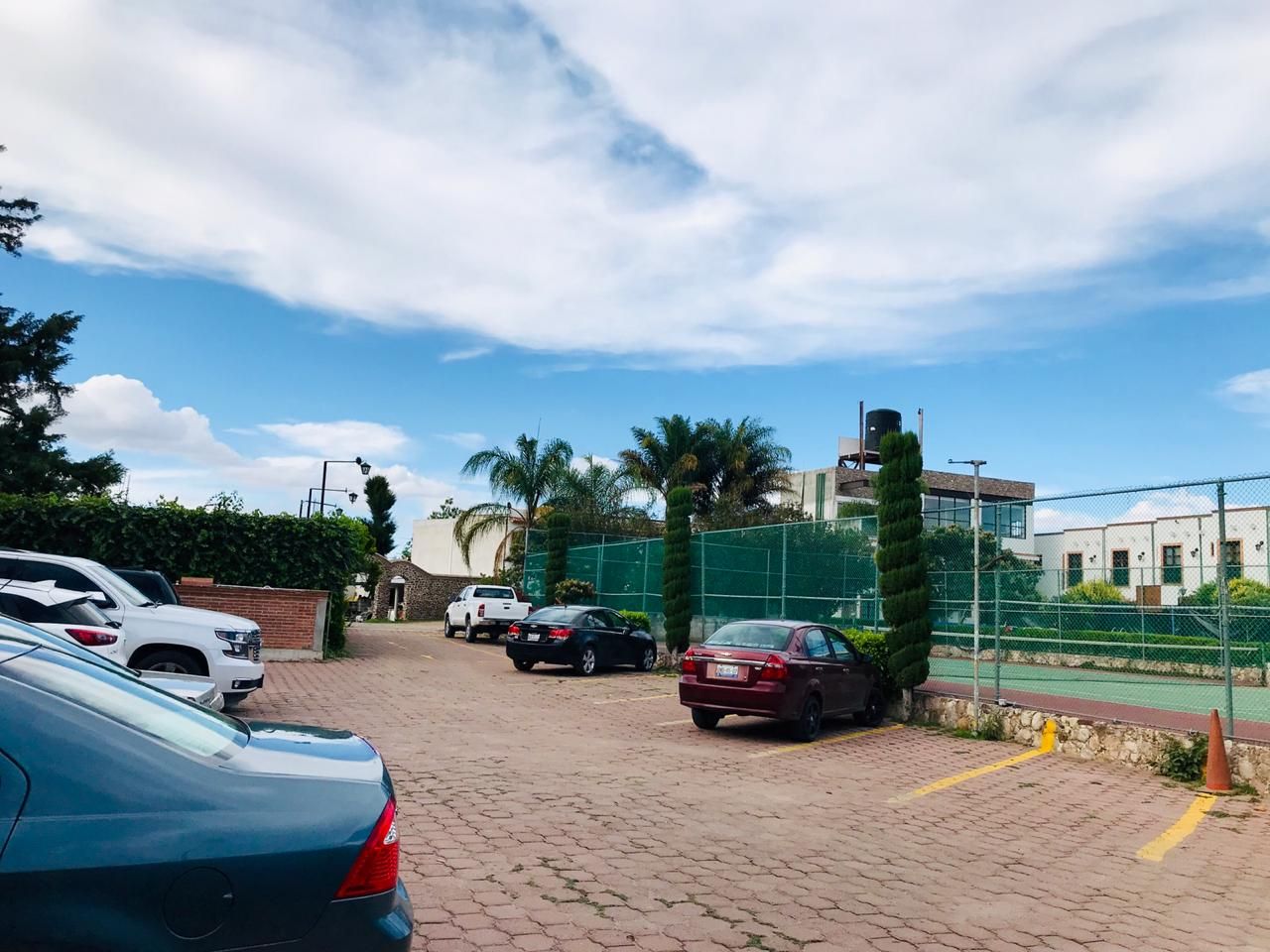 Estacionamiento con varios autos junto a una cancha deportiva cercada bajo un cielo parcialmente nublado.