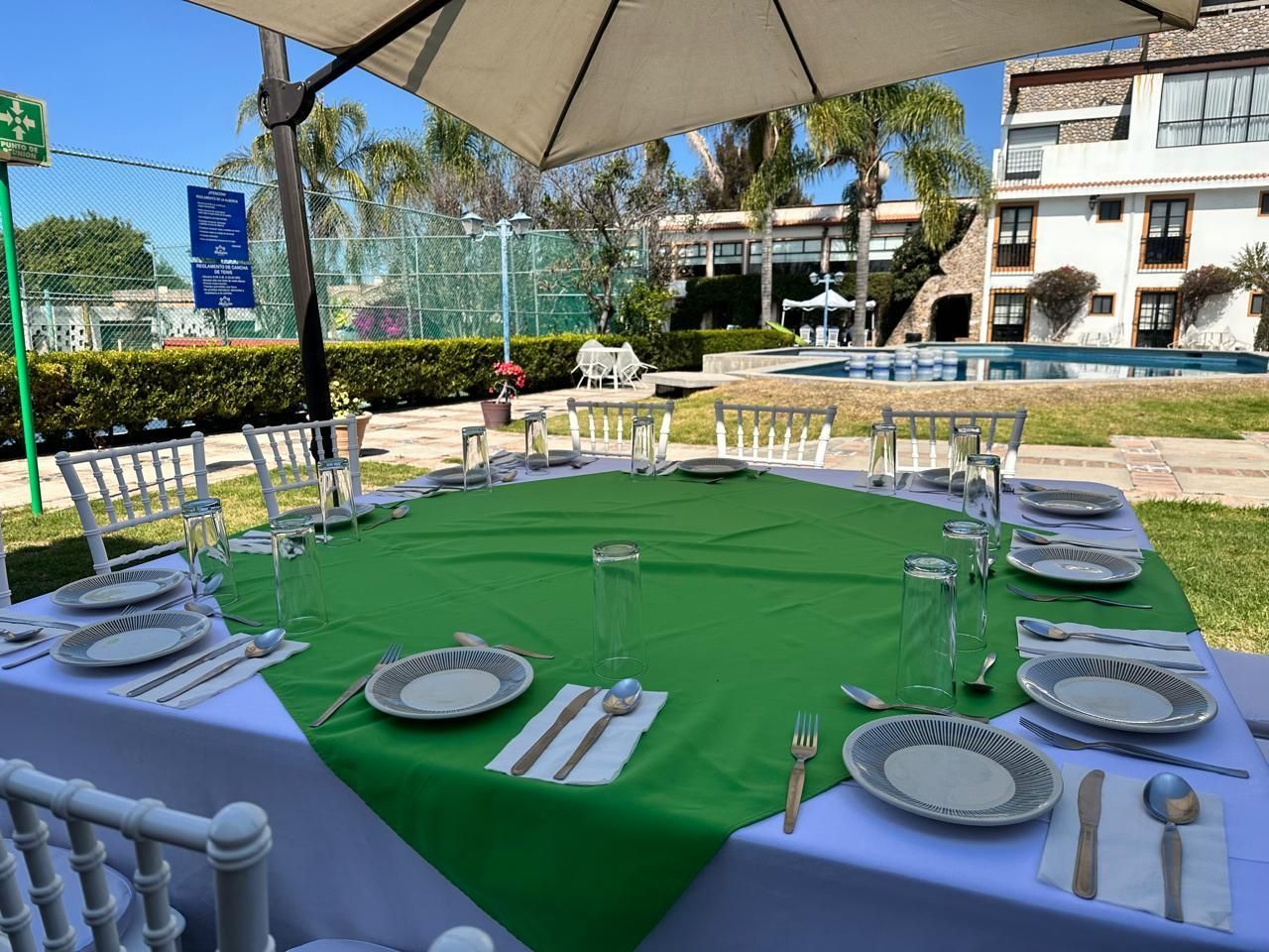 Juego de mesa y sillas de exterior con mantel verde, sillas blancas y platos bajo una sombrilla de patio junto a la piscina.