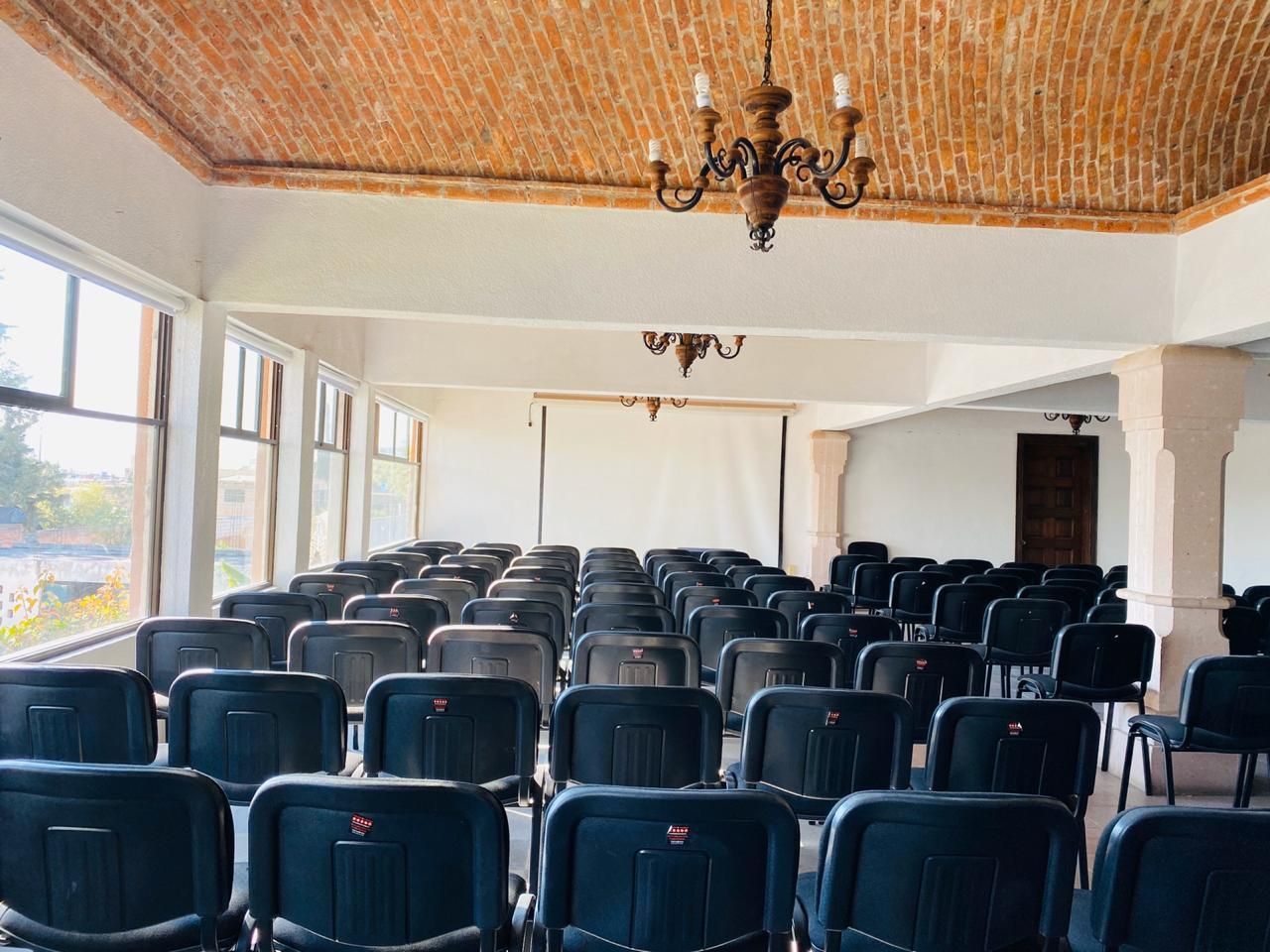 Auditorio vacío con filas de asientos azul oscuro, candelabros y grandes ventanales que dejan entrar la luz del día.