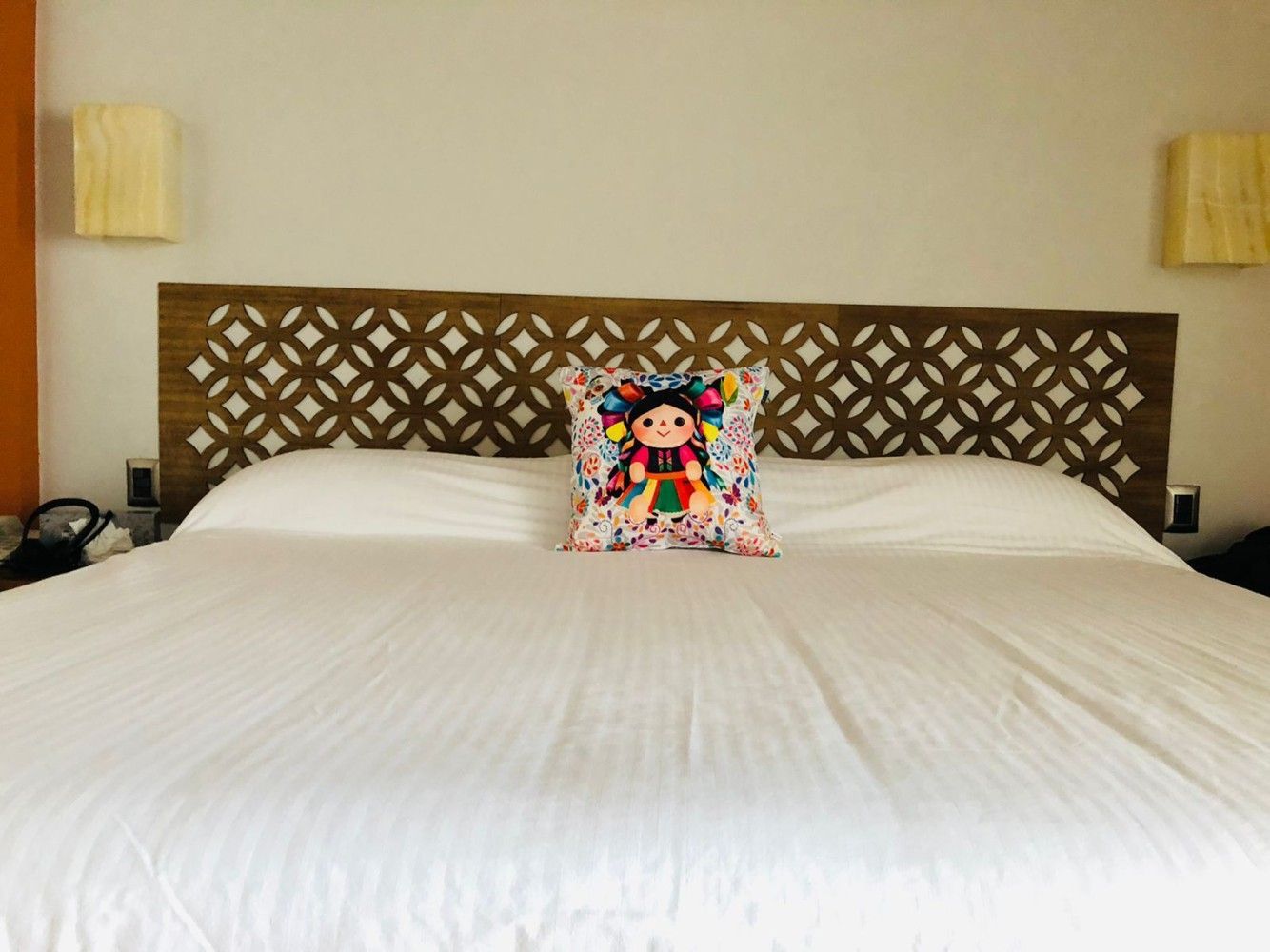 Cama de hotel con sábanas blancas, cabecero estampado y un cojín decorativo colorido centrado entre las almohadas.