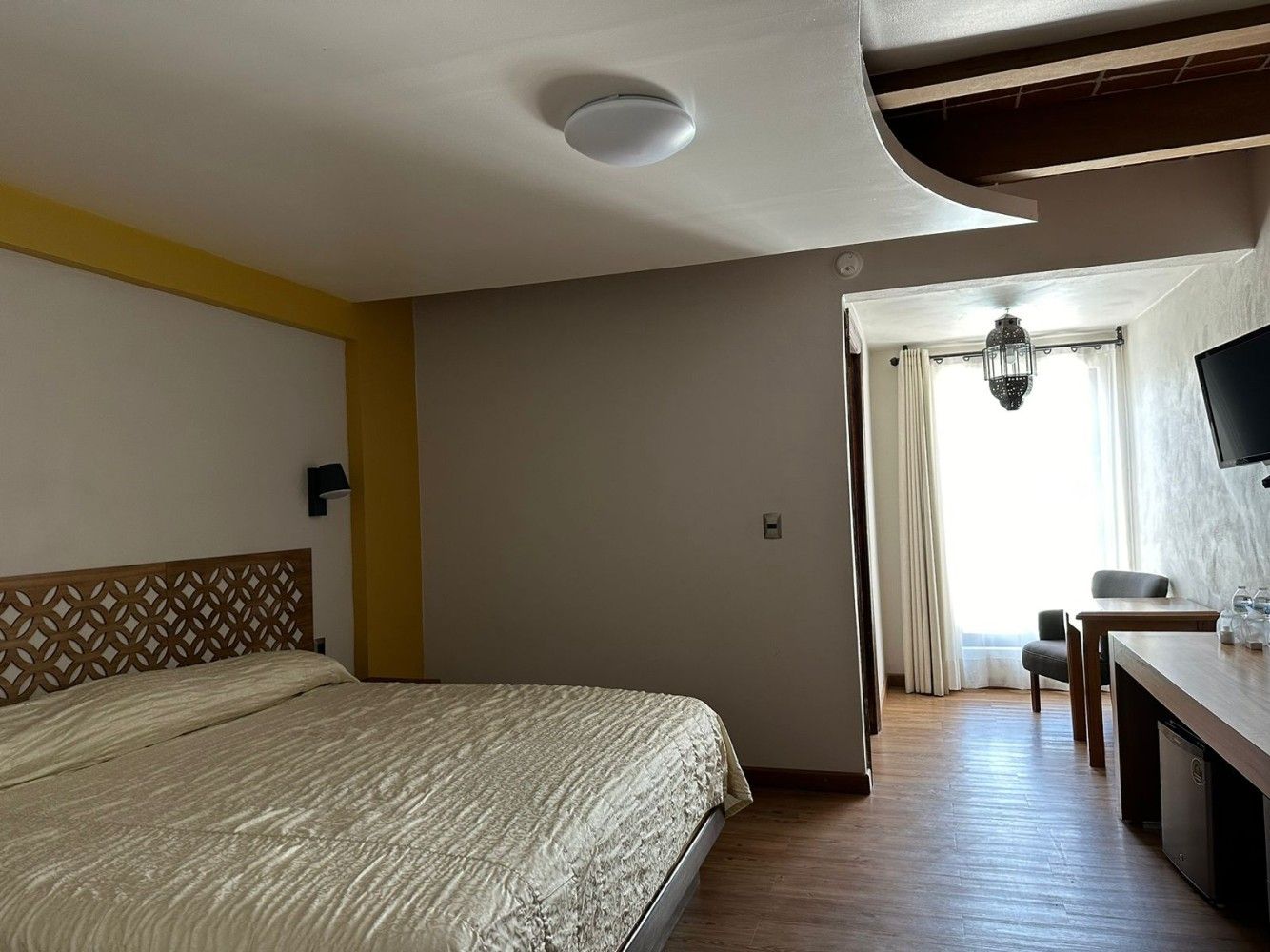 Habitación con cama doble, paredes de color beige, suelo de madera y una luminosa zona de estar junto a la ventana.