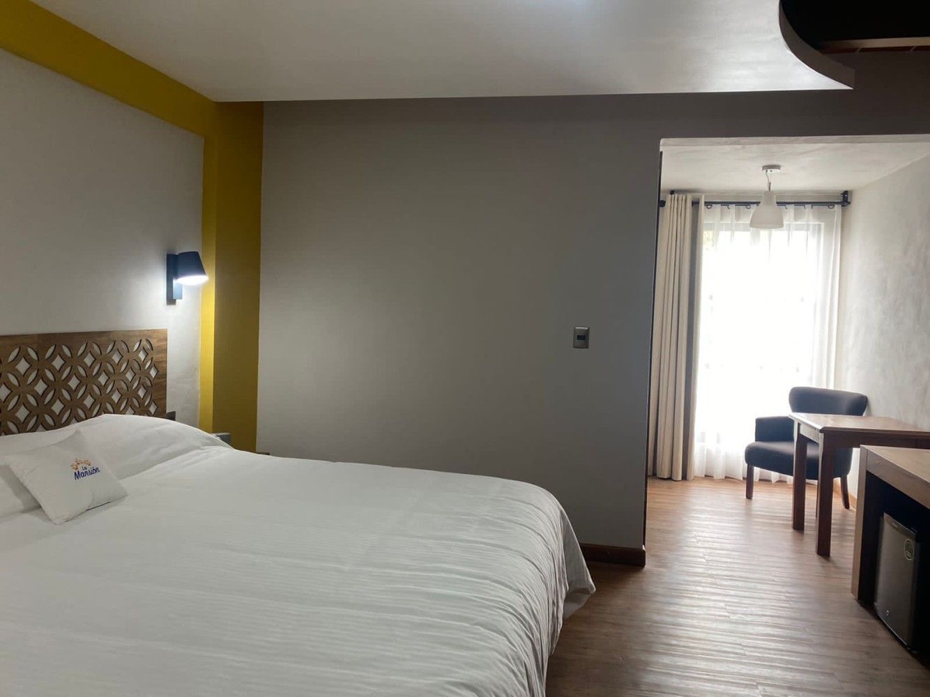 Habitación de hotel moderna con cama hecha, pared decorativa amarilla, escritorio y zona de estar junto a la ventana.