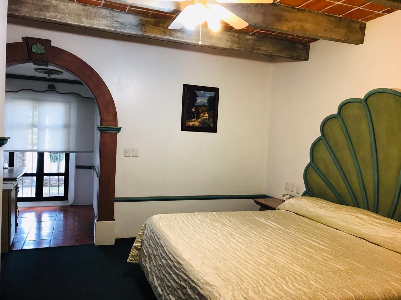Dormitorio con vigas de madera en el techo, puerta arqueada y cama con cabecero festoneado de color verde.