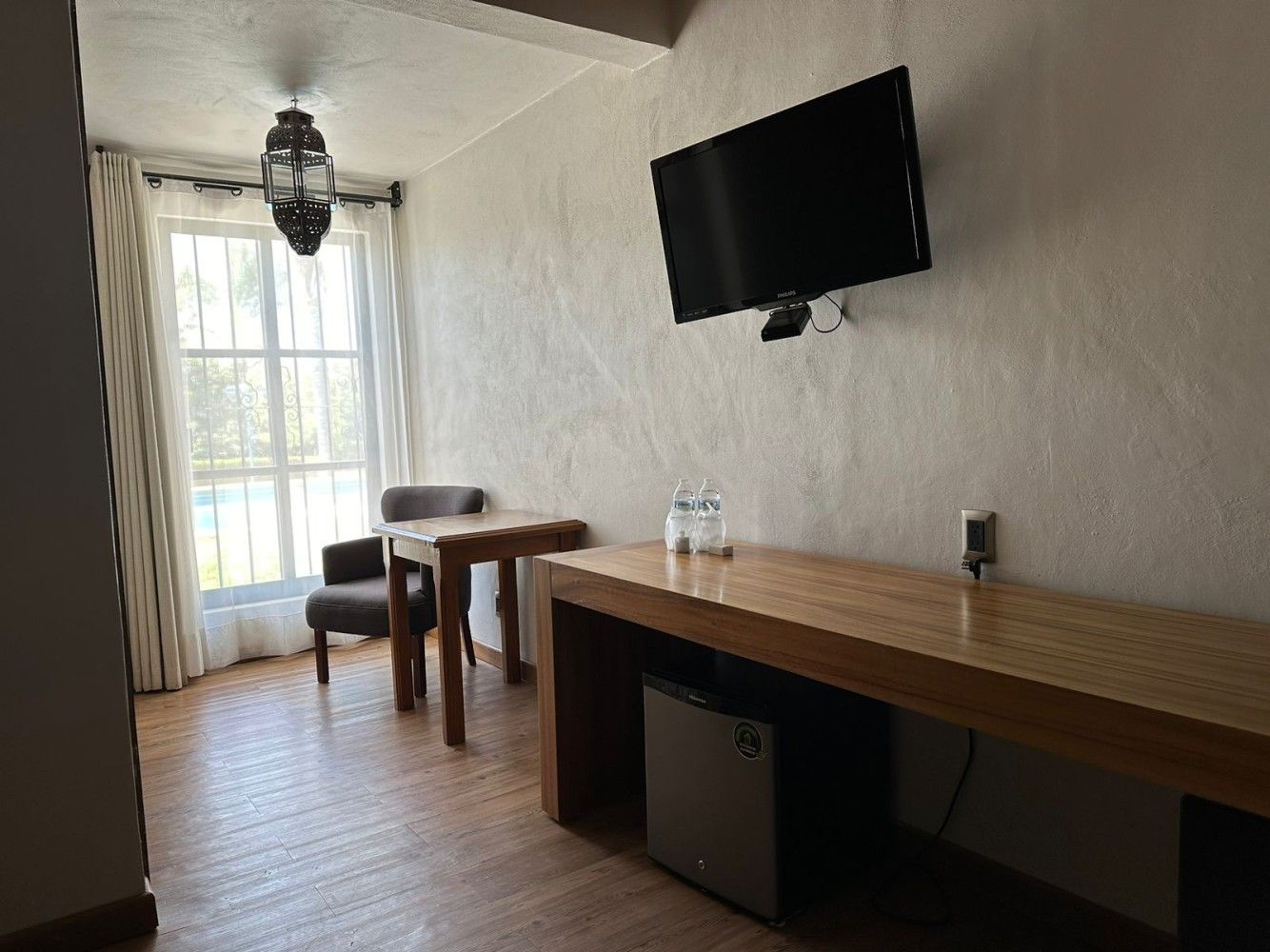 Una pequeña habitación de hotel con televisor de pared, escritorio, silla, ventana y mininevera.