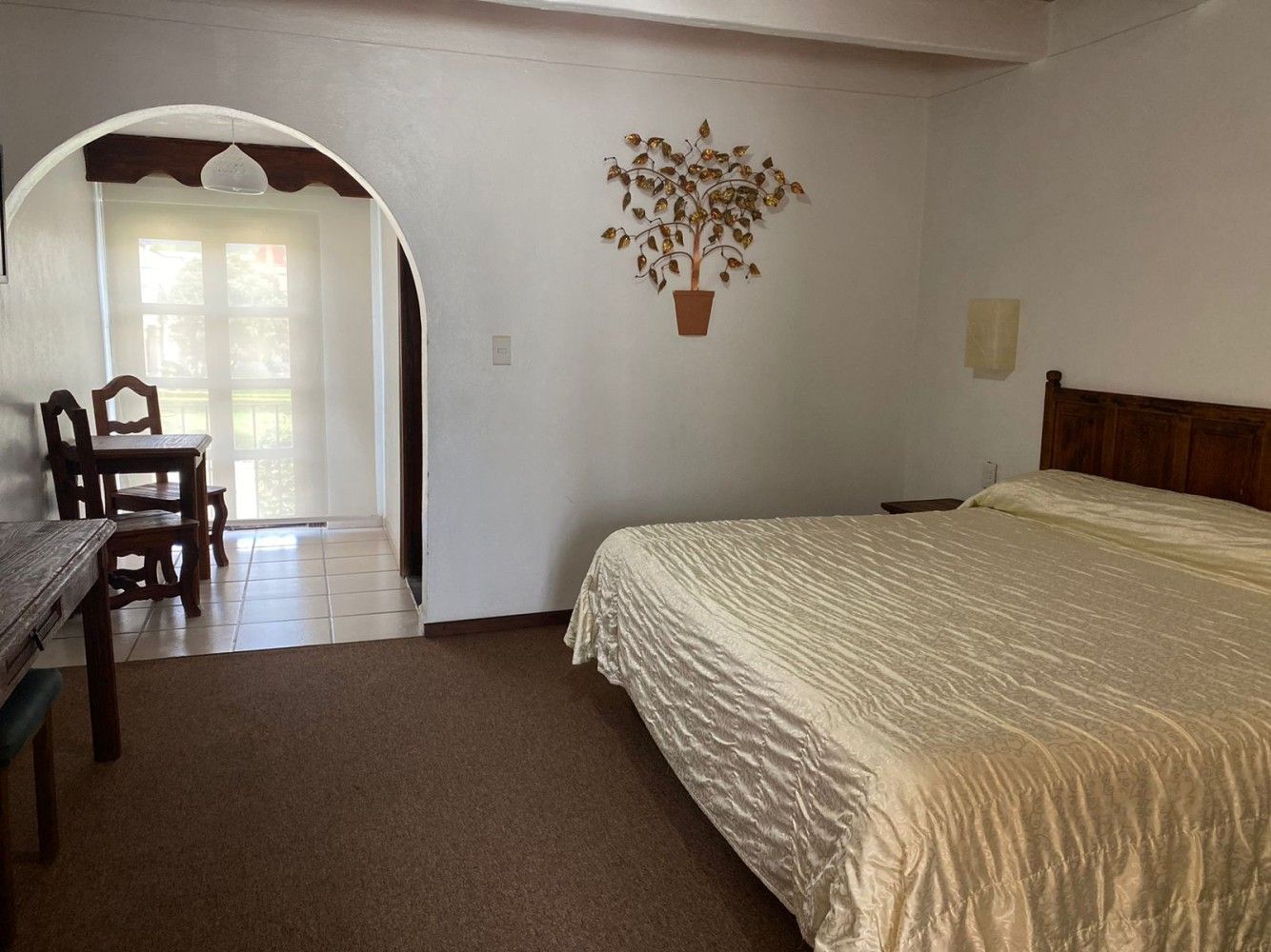 Habitación con cama, moqueta marrón, puerta arqueada y mesa junto a una ventana luminosa.
