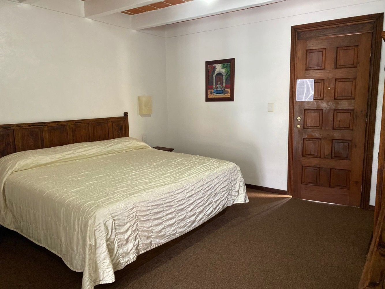 Habitación sencilla con una cama, puerta de madera y un pequeño cuadro enmarcado en una pared blanca.