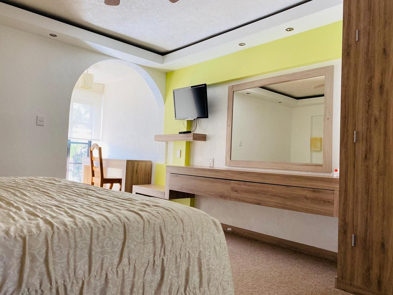 Habitación luminosa con cama, ventana arqueada, espejo de pared, televisor y armario de madera.