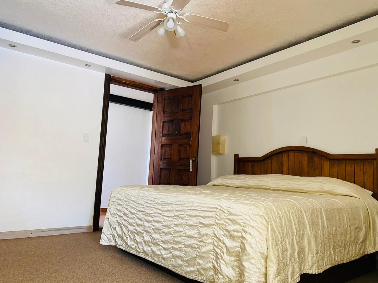 Dormitorio con una cama grande, cabecero de madera, puerta abierta y moqueta beige sobre techo blanco.