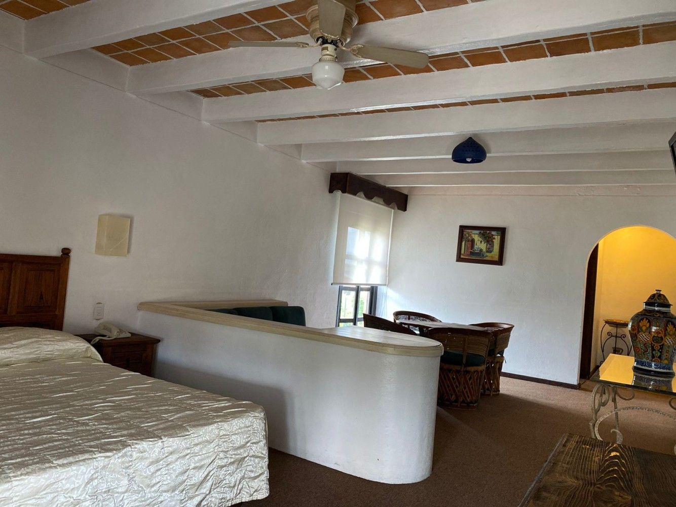 Dormitorio con paredes blancas, techo con vigas de madera a la vista, cama y puerta arqueada que da a una habitación contigua.