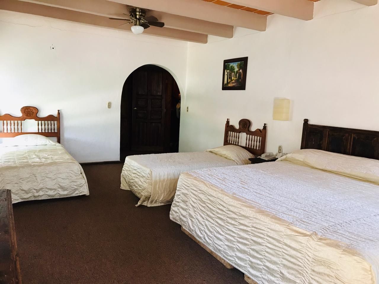 Habitación espaciosa con tres camas, paredes blancas, vigas vistas y una puerta arqueada oscura.