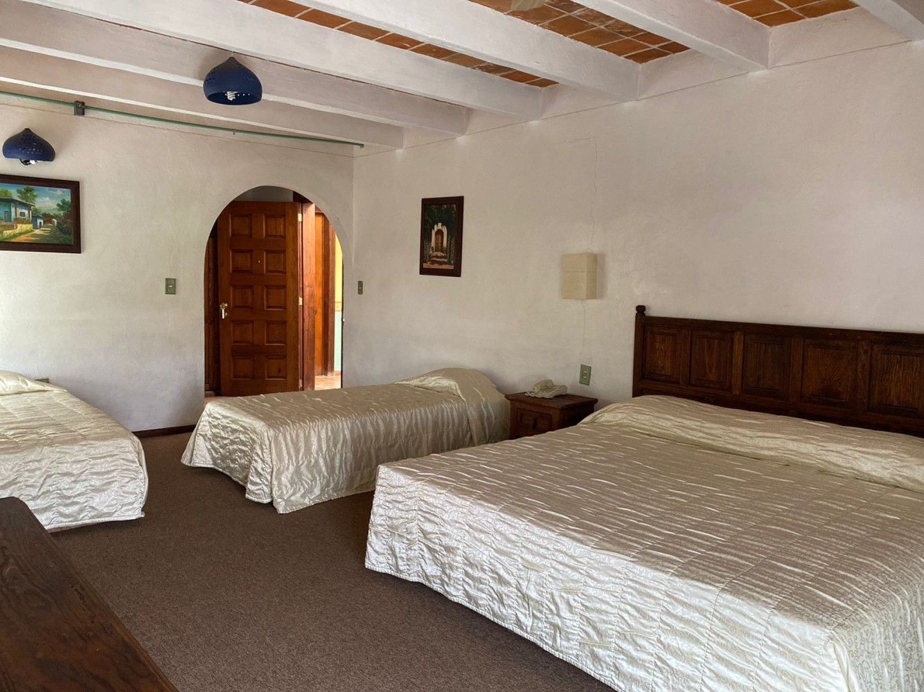 Tres camas en una habitación rústica con paredes blancas, vigas a la vista y una puerta de madera arqueada.