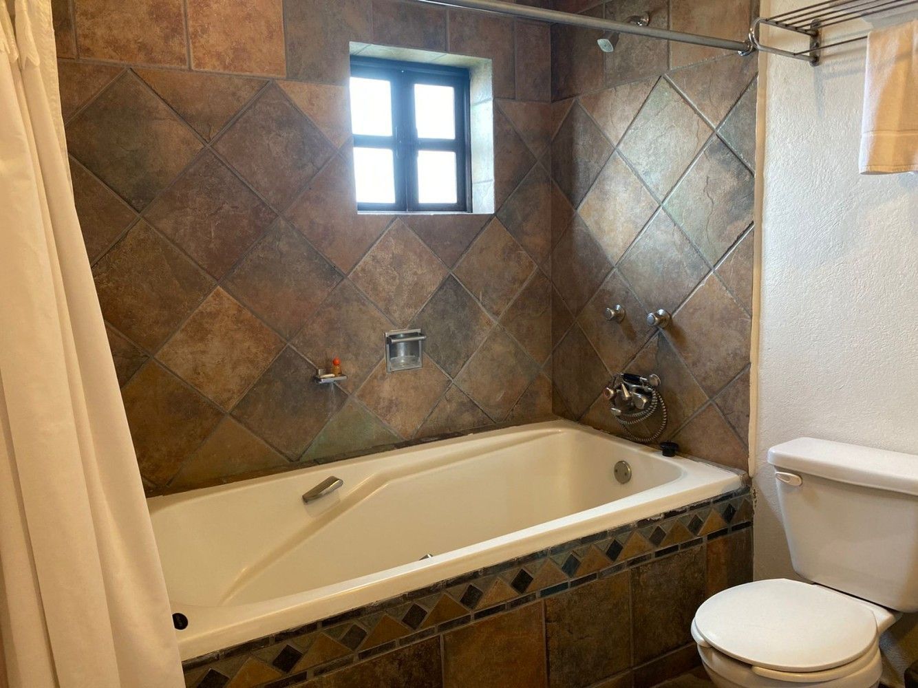 Baño con bañera blanca, paredes alicatadas, ventana, inodoro y cortina de ducha beige.